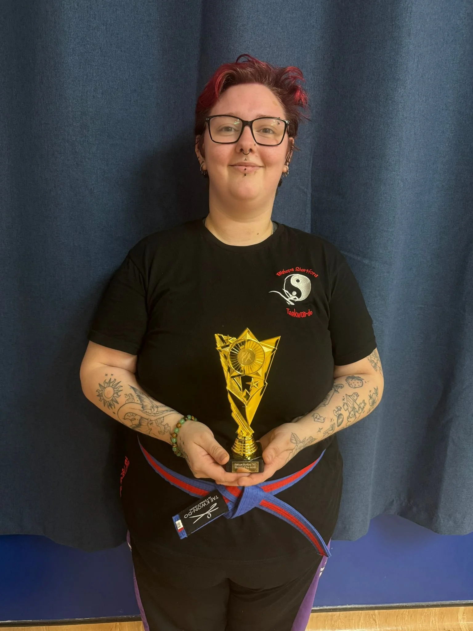 Person with tattoos on arms and glasses holding a gold trophy while standing in front of a blue curtain.