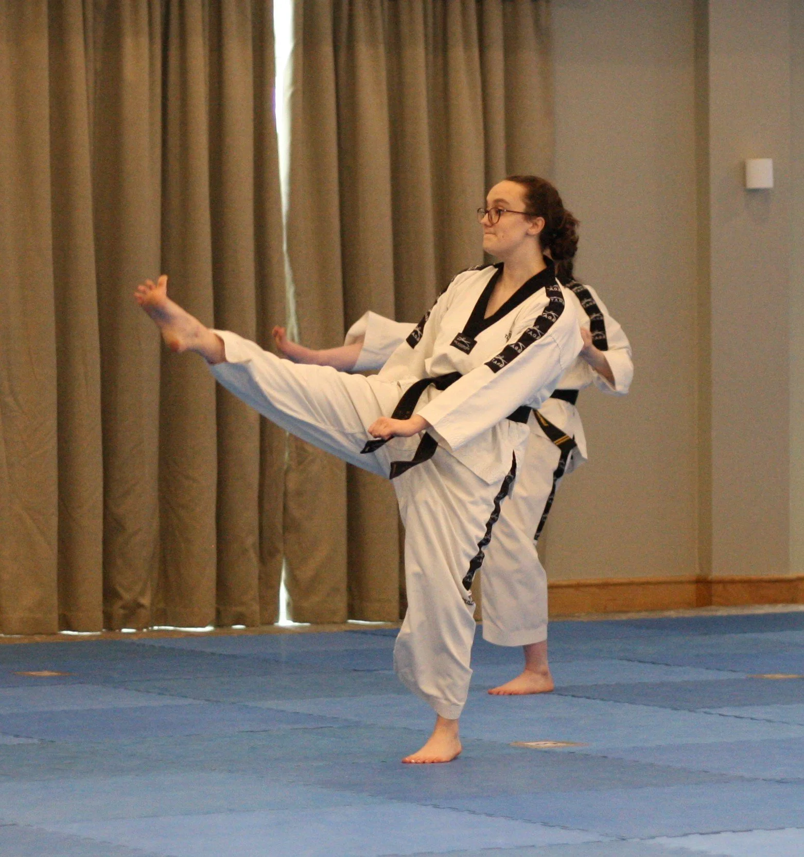 Two martial artists practicing martial arts on a blue mat indoor, with beige curtains in the background. The person in the foreground is executing a high kick, wearing a white gi with black trim and a black belt. The person in the background is also 