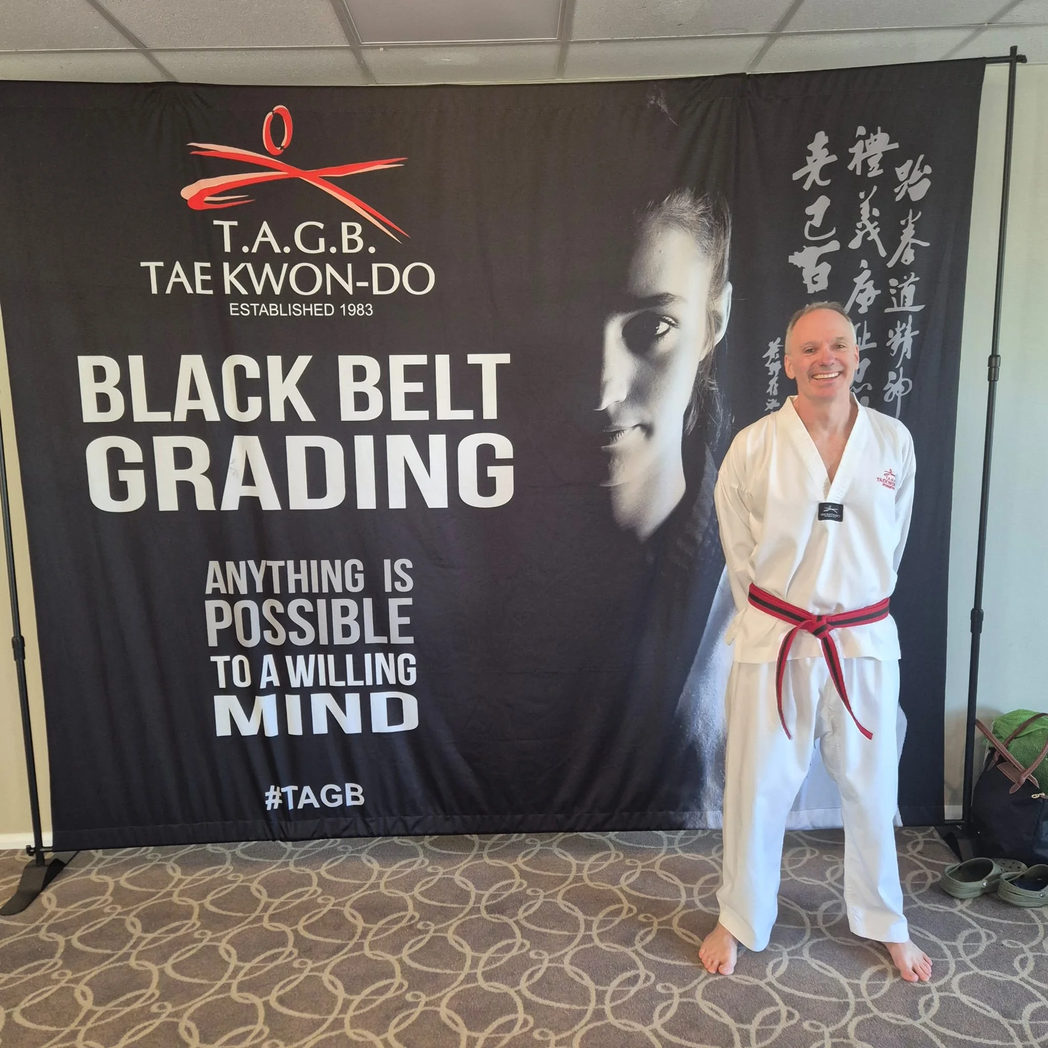 A smiling man in a martial arts uniform with a black belt standing in front of a banner that reads "T.A.G.B. Tae Kwon-Do established 1983" and "Black Belt Grading" with Asian characters on the right side.