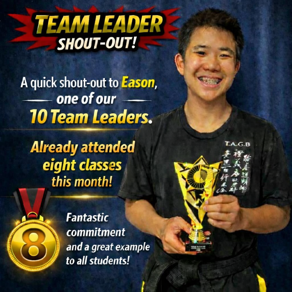 A young man wearing a black martial arts uniform holding a gold trophy, smiling. The background features a blue curtain with text recognizing him as a team leader and praising his dedication in martial arts classes.