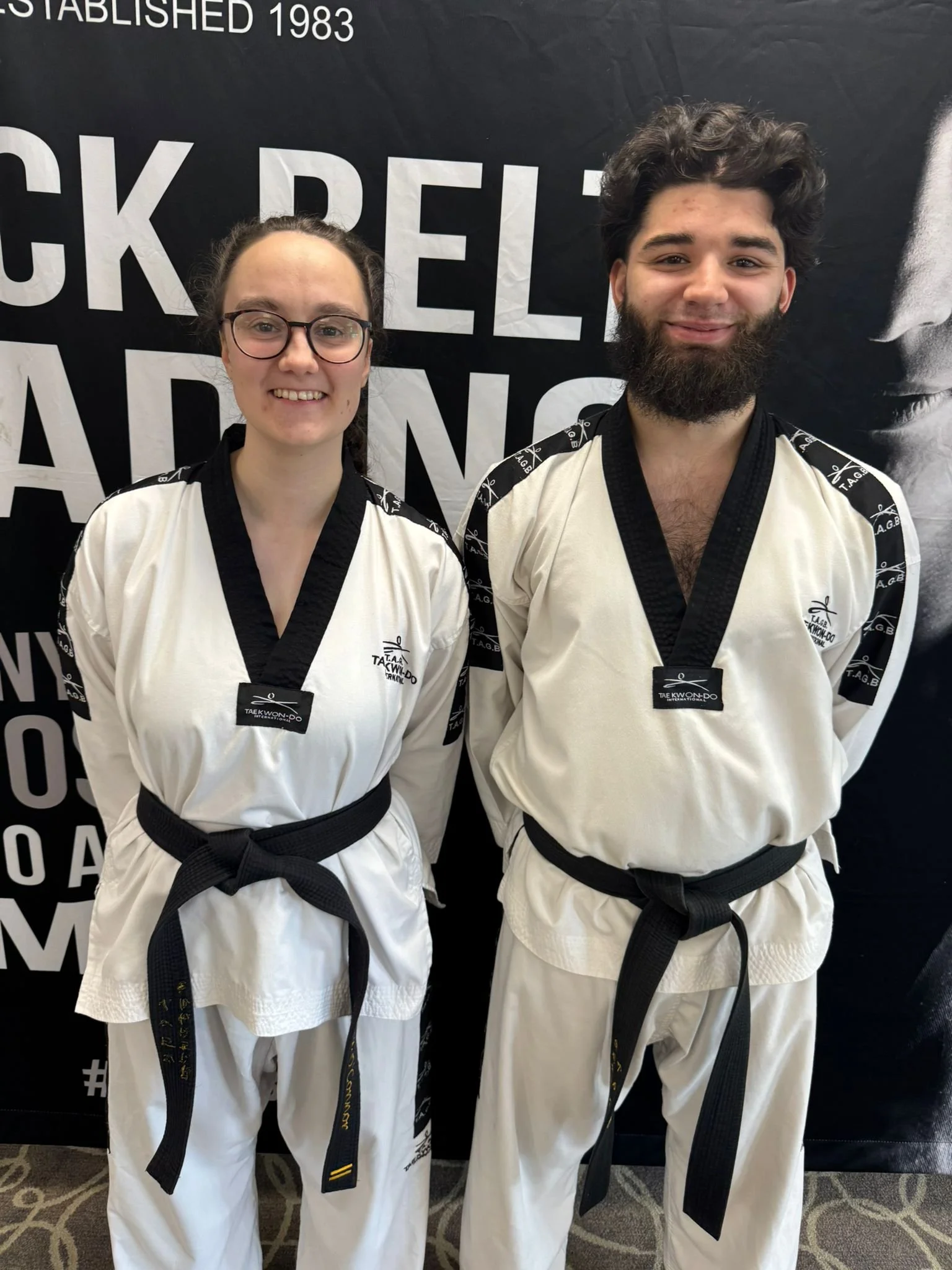 Two young martial artists wearing white uniforms and black belts, posing together indoors against a black backdrop with white text, smiling at the camera.