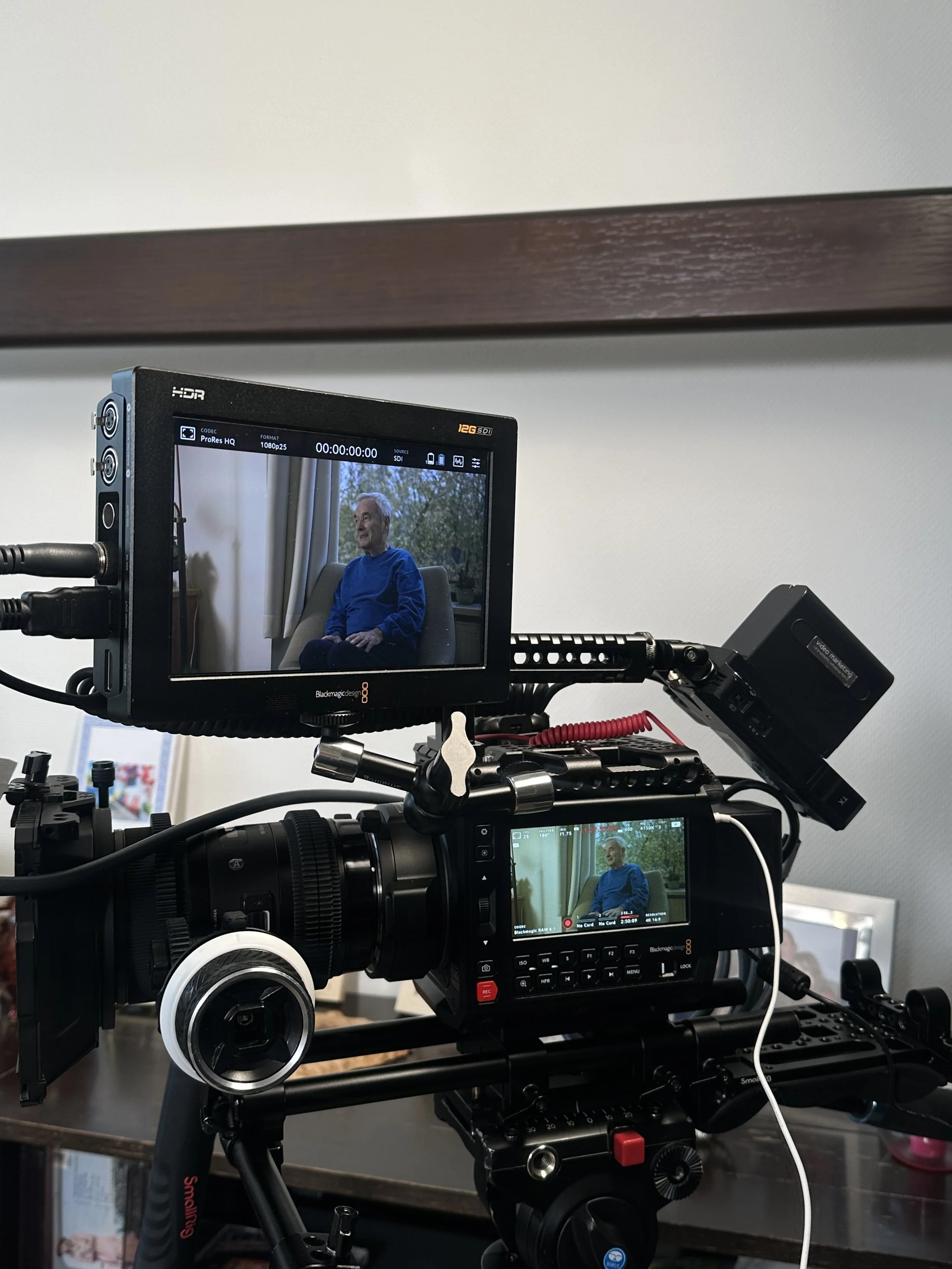 Camera equipment set up on a tripod with a monitor displaying a man sitting in a chair, wearing a blue shirt and smiling, with a window and curtains in the background.