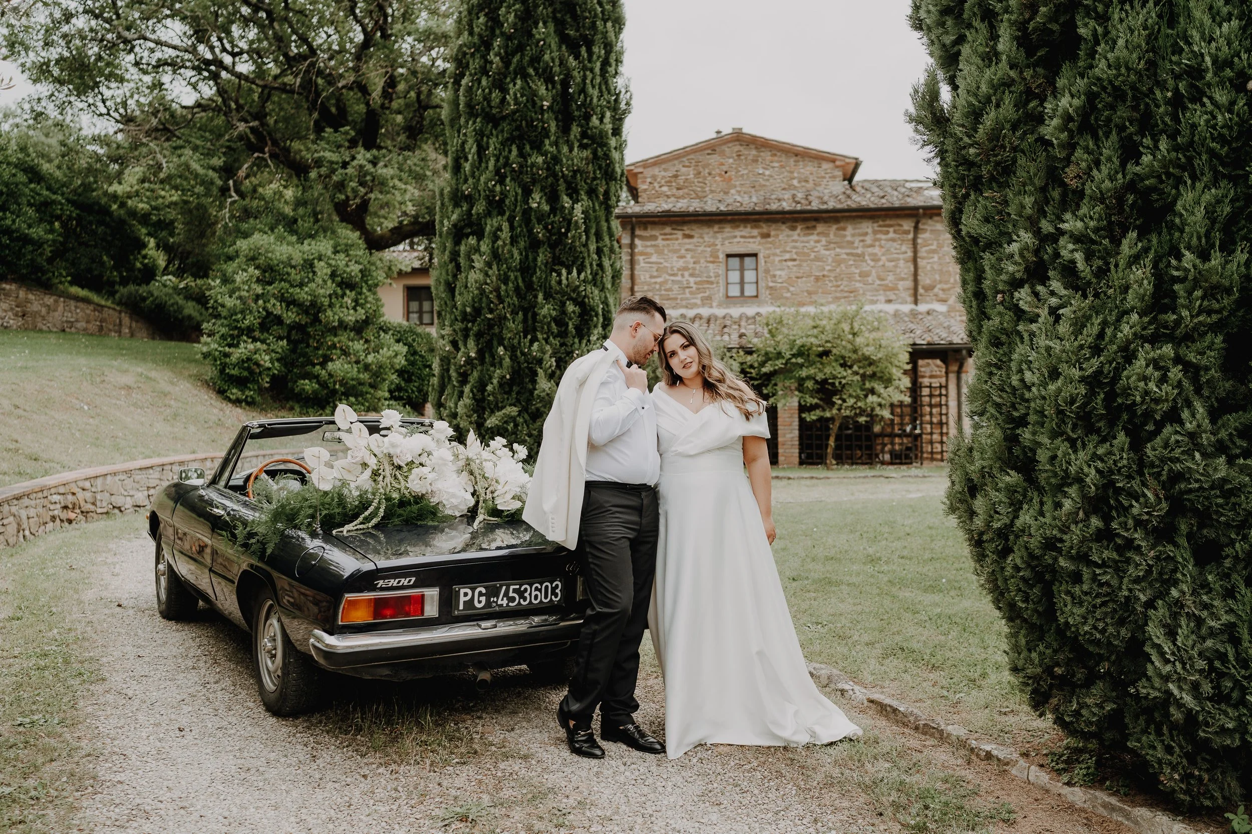 Brautpaar umarmt sich auf einem Kiesweg, Cabrio im Hintergrund, Bäume und Zypressen – Hochzeitsreportage Toskana, Italien. Fotografin Claudia Hilgenböker aus Lemgo - Destination Wedding