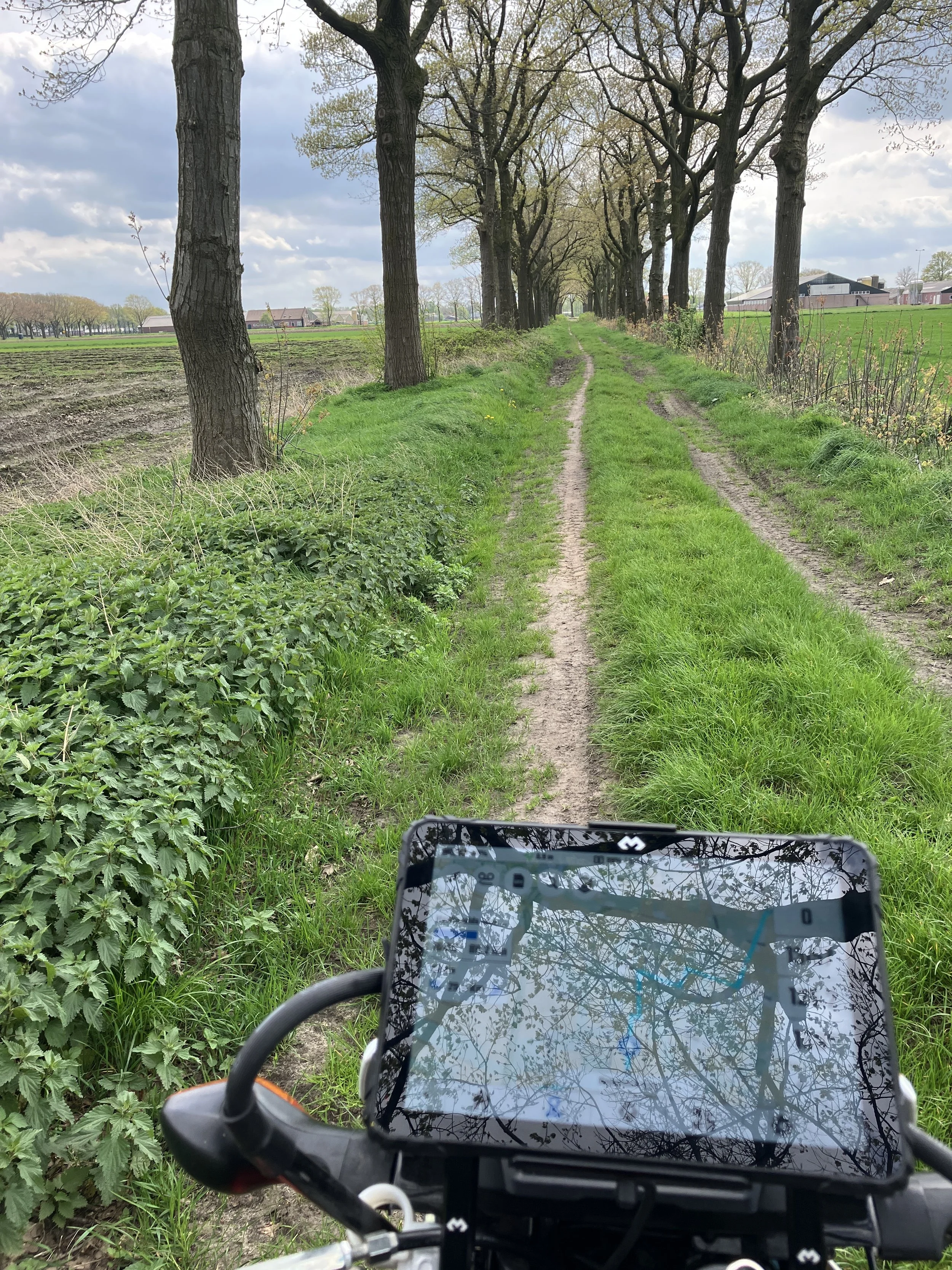 Allroad Motor Guide op een onverharde landweg met sporen van voertuigen, omzoomd door bomen en groene velden onder een bewolkte hemel. Op de voorgrond is een allroad motor te zien met een DMD2-apparaat bevestigd.
