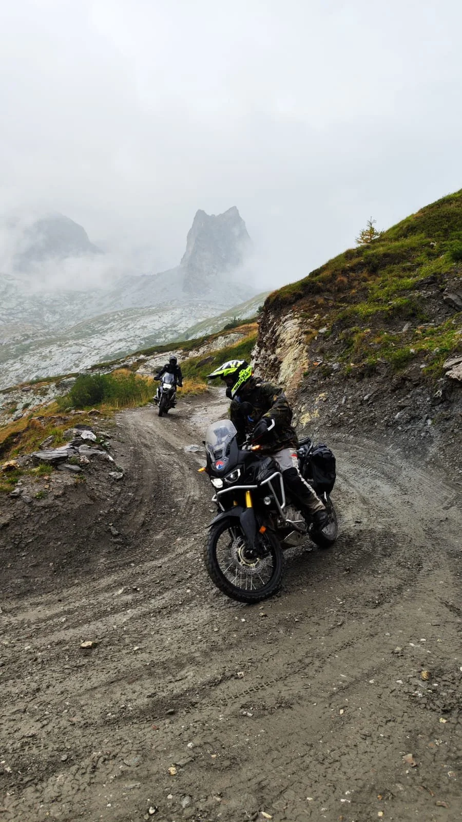 Twee allroad motoren rijden over een modderige bergweg in een bergachtig landschap, dicht bij een rotsformatie en mistige heuvels op de achtergrond.