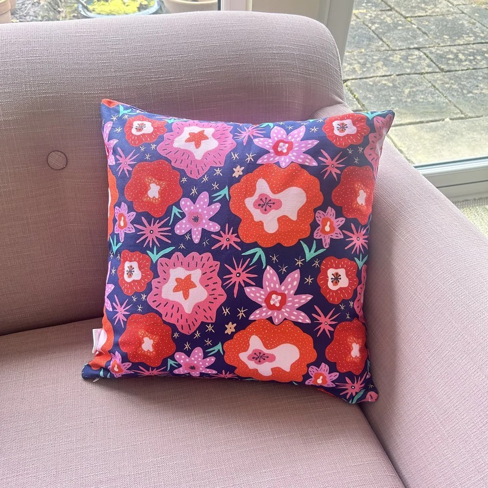 Colourful floral patterned cushion featuring red pink orange and lilac flowers by Jennie Fynn on a pink sofa