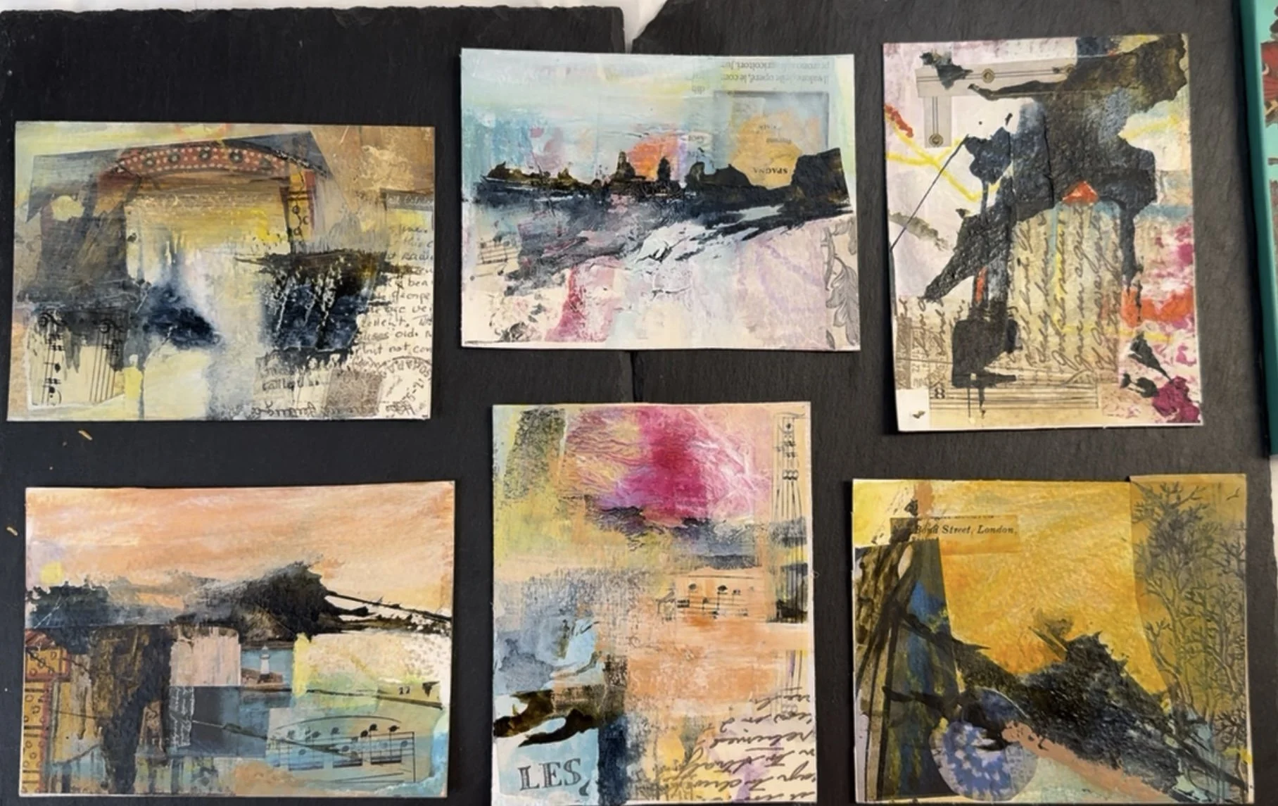 Six mixed media art postcards with abstract landscapes, handwritten script, and musical notes, arranged on a black surface.