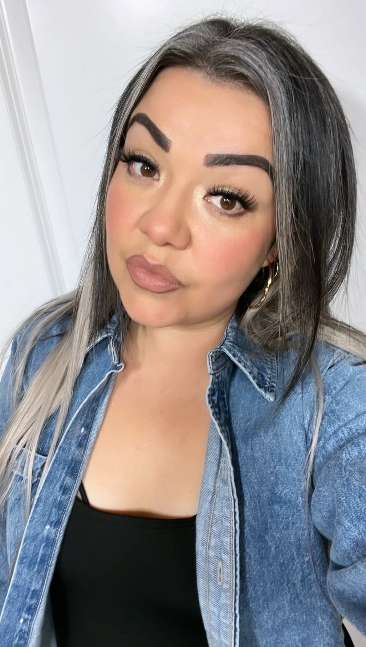 A woman with long, dark hair with gray streaks, wearing a denim shirt over a black top, posing indoors with a neutral expression.