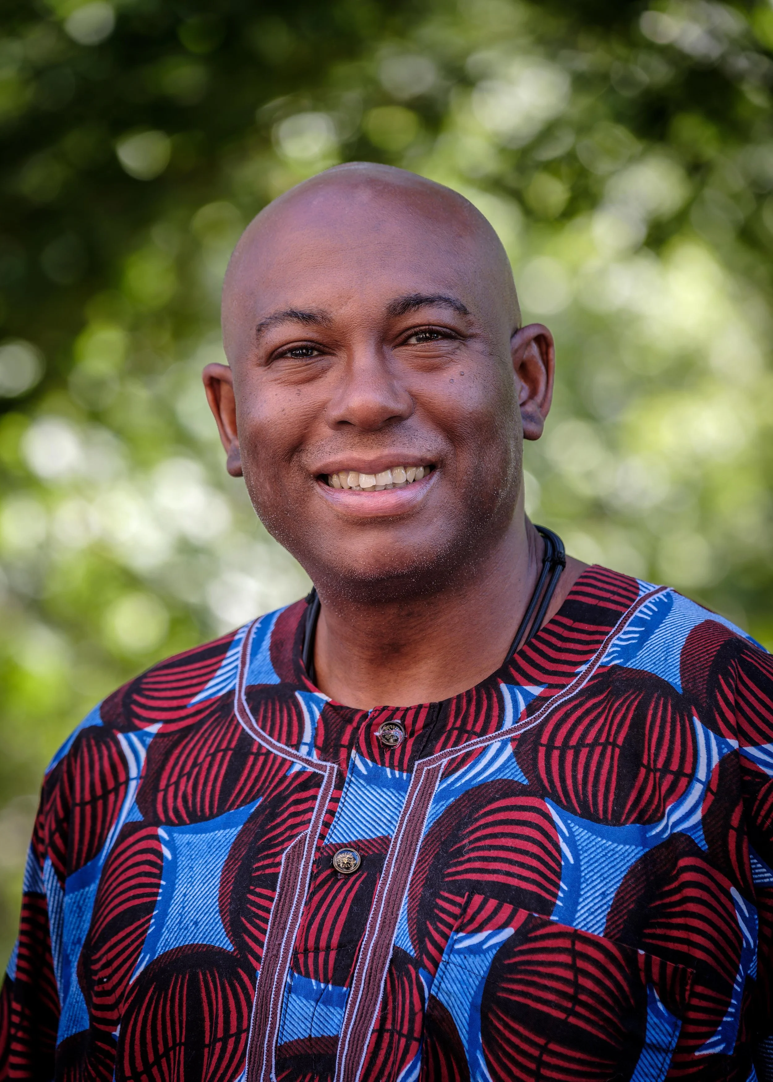 A smiling man with a shaved head wearing a colorful patterned shirt outdoors with a blurred green background.