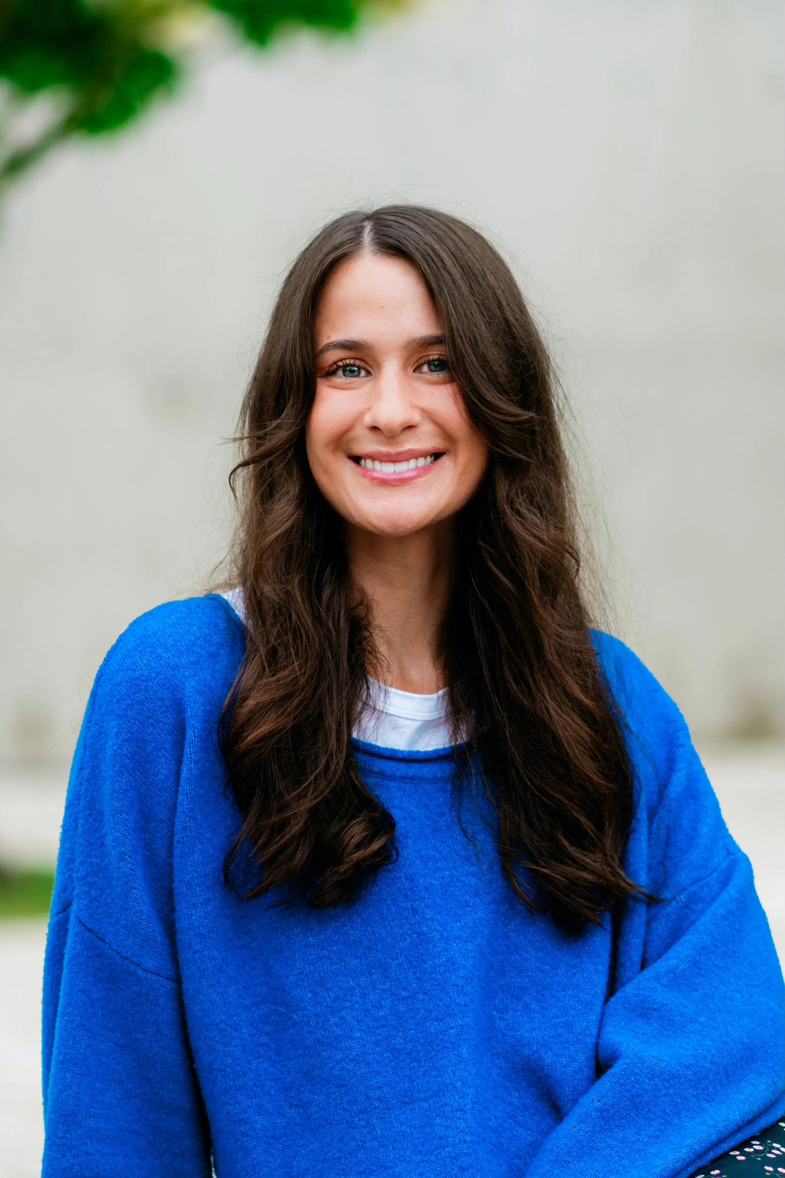A smiling young woman with brown wavy hair wearing a tan coat and gray sweater, standing outdoors in a park with autumn leaves, trees, picnic tables, and people in the background.