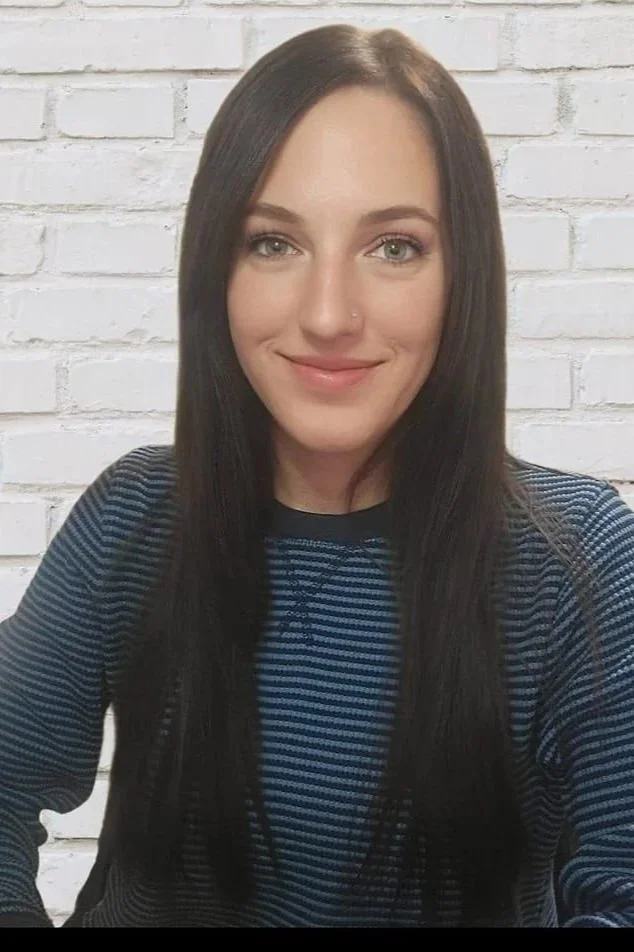 A young woman with long dark hair and blue eyes, smiling, wearing a blue and black striped shirt, sitting against a white brick wall.