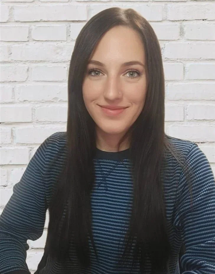A woman with long dark hair, fair skin, and blue eyes, smiling in front of a white brick wall, wearing a blue and black striped long-sleeve shirt.