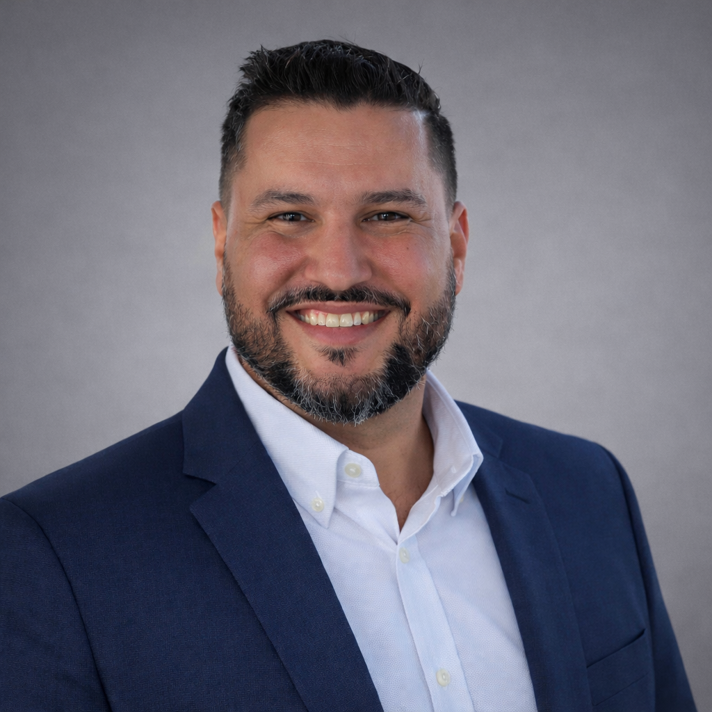 A smiling man with dark hair and a beard wearing a white shirt stands on a beach with the ocean and blue sky in the background.