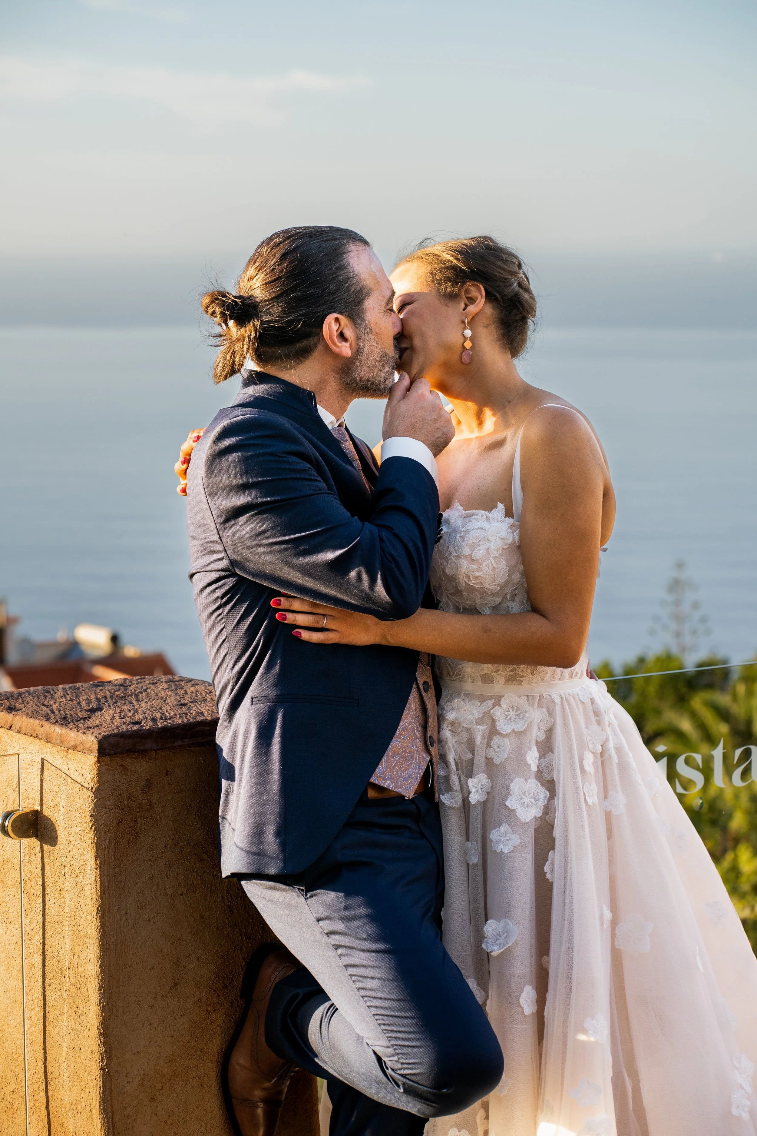 Ein Mann und eine Frau küssen sich bei Sonnenuntergang an einer Wasserfront, mit Blick auf das Meer im Hintergrund.