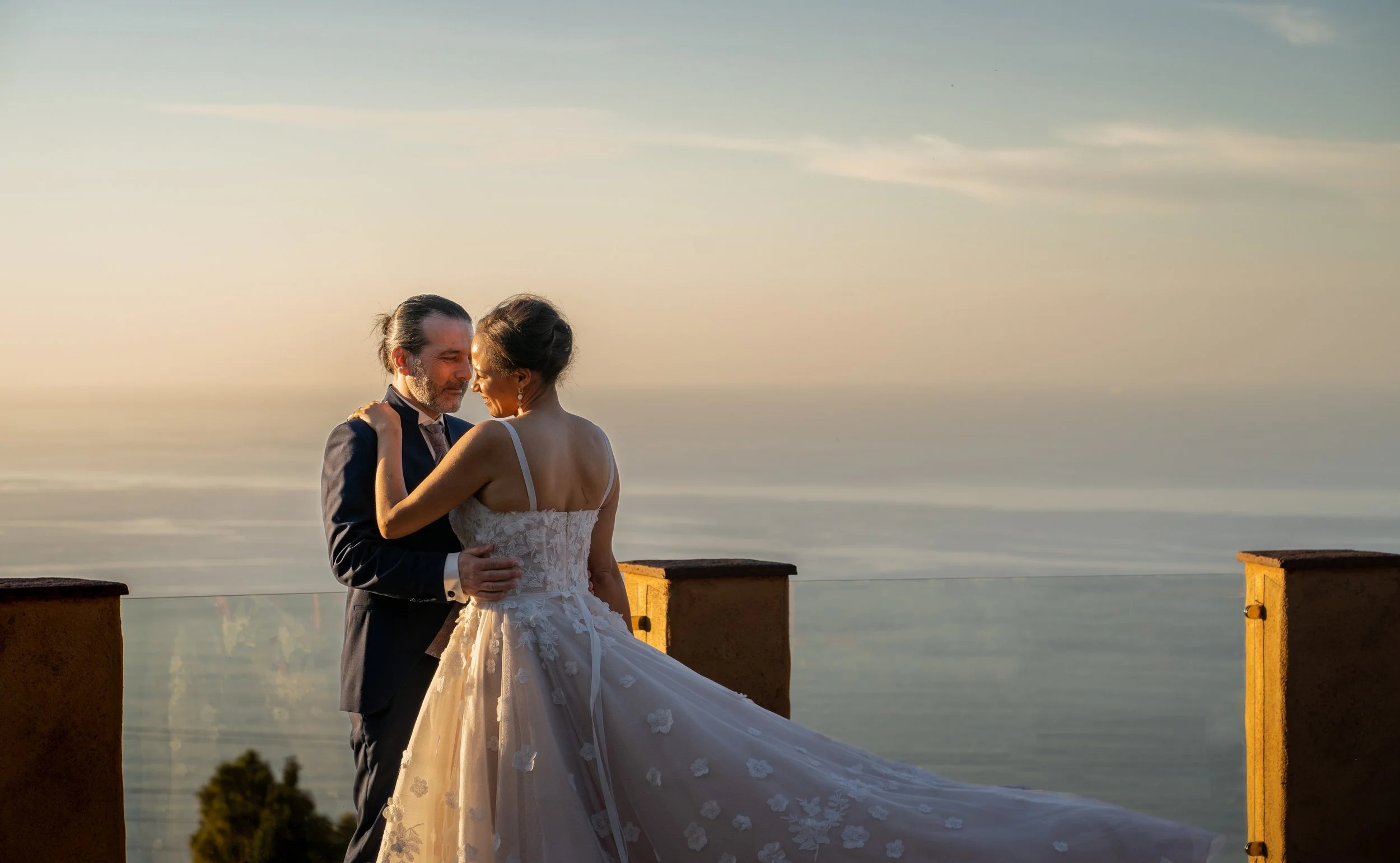 Ein gut gekleideter Mann und eine Braut, die sich beispielsweise bei einer Hochzeit am Meer umarmen, während sie auf einem Balkon vor dem Wasser stehen, bei Sonnenuntergang oder Dämmerung.