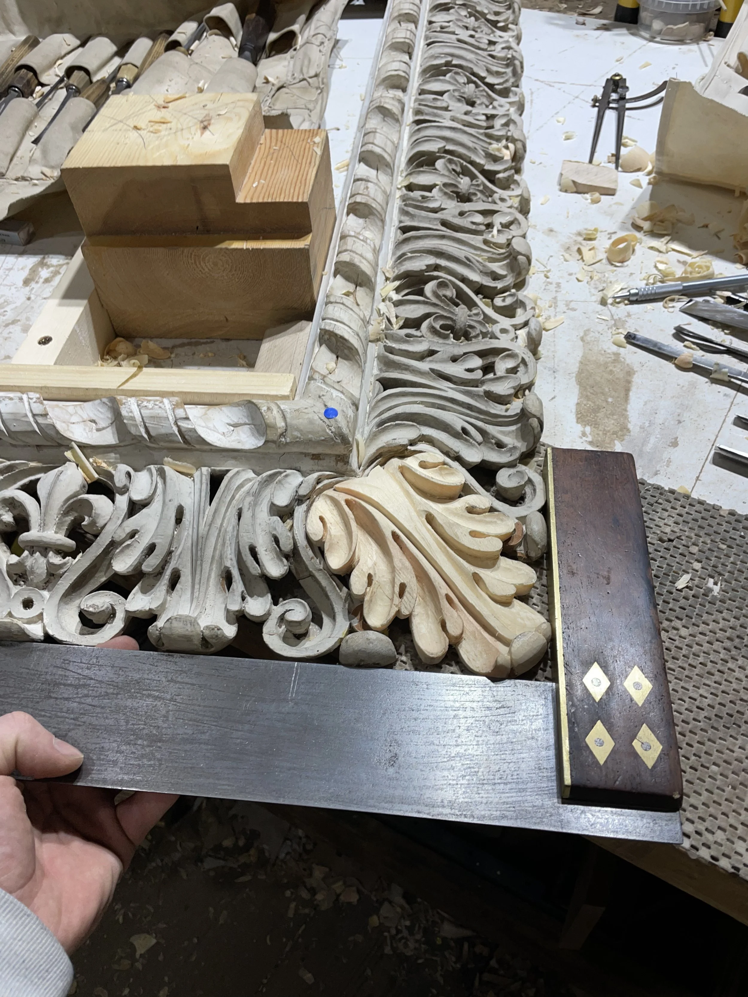 Close-up of detailed wood carving work in progress with ornamental scrolls and decorative elements, surrounded by woodworking tools and wood shavings.