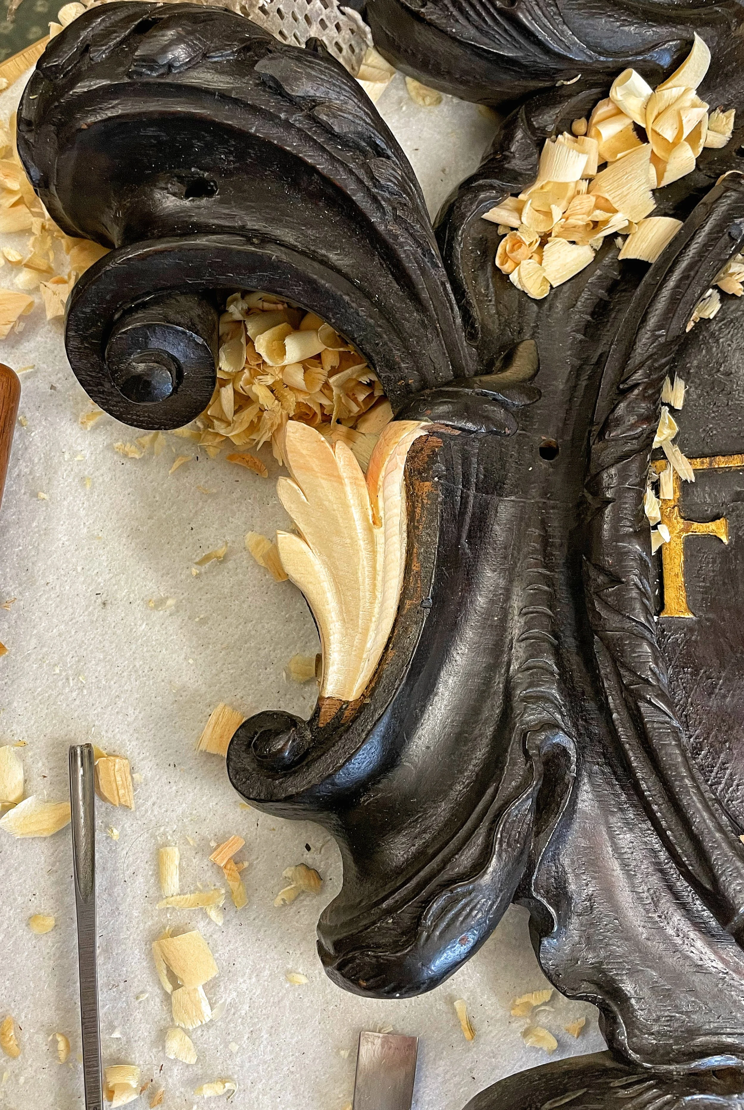 Close-up of an ornate wooden carving in progress, with wood shavings scattered around, showing black and light-colored wood details.