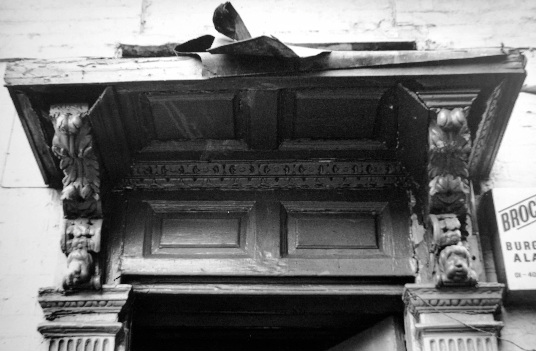 An ornate, antique wooden cabinet with elaborate carvings on the corners, situated above a wooden panel, with a couple of small objects on top and a partially visible sign in the background.