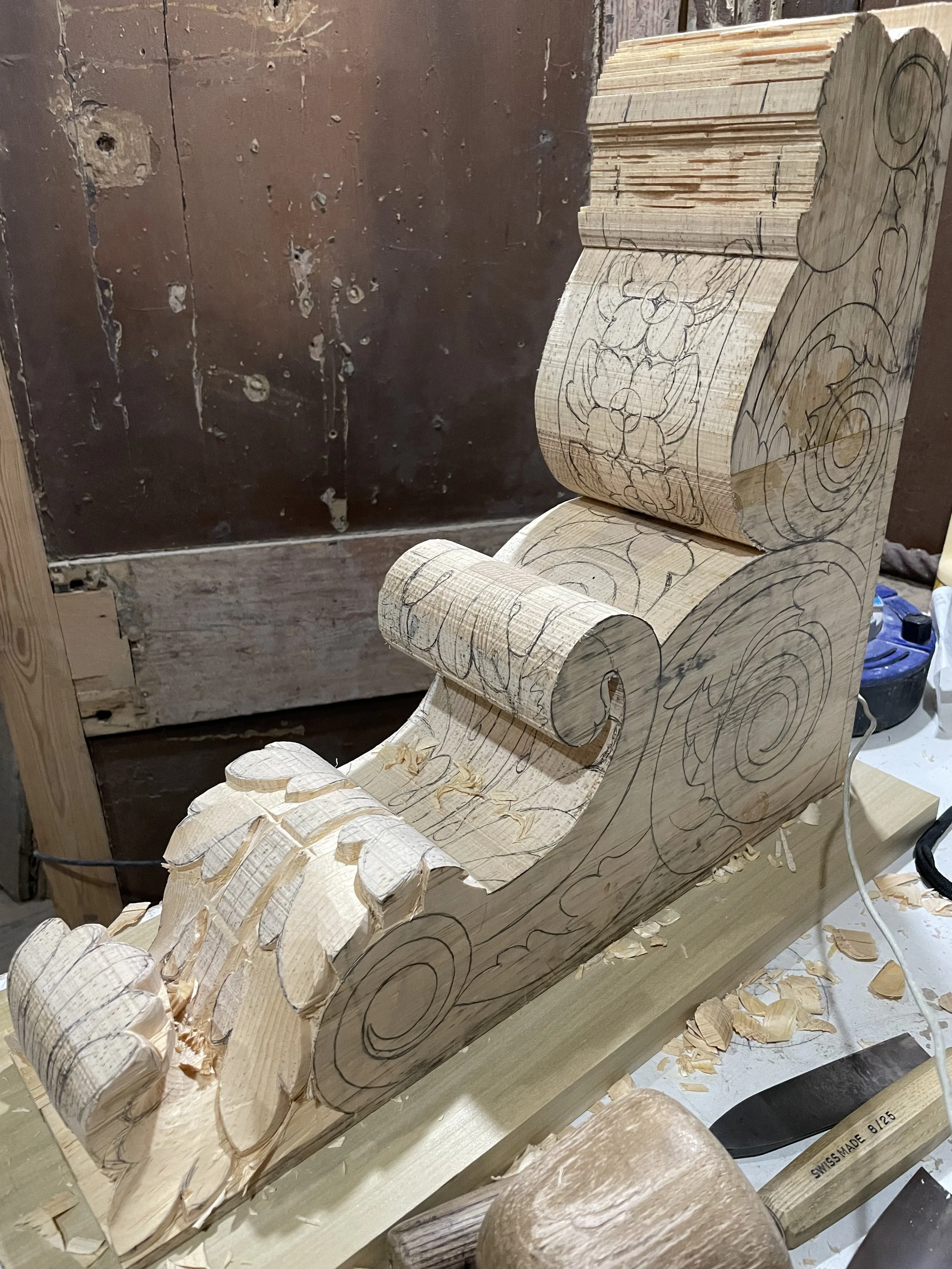 A work-in-progress ornate wood carving with detailed swirl and floral patterns, partially shaped and marked with pencil lines, sitting on a workbench in a woodworking shop.