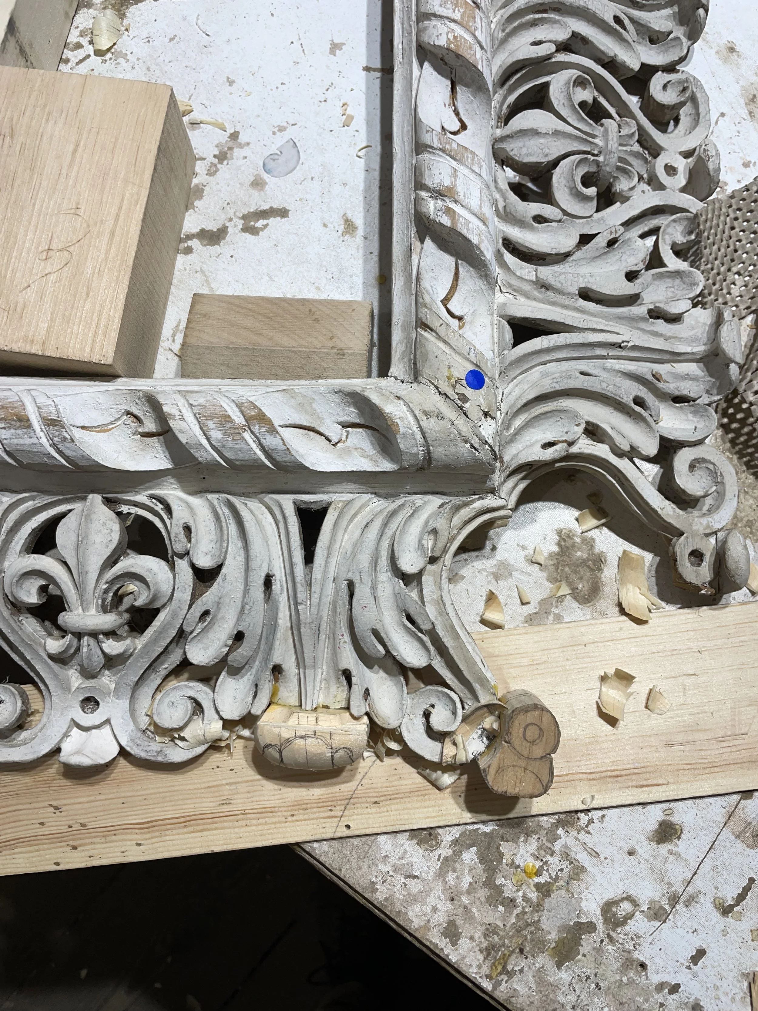 Close-up of a decorative, ornate, white-painted wooden corner trim with elaborate scroll and floral carvings, partially repaired and surrounded by wood shavings and construction materials.