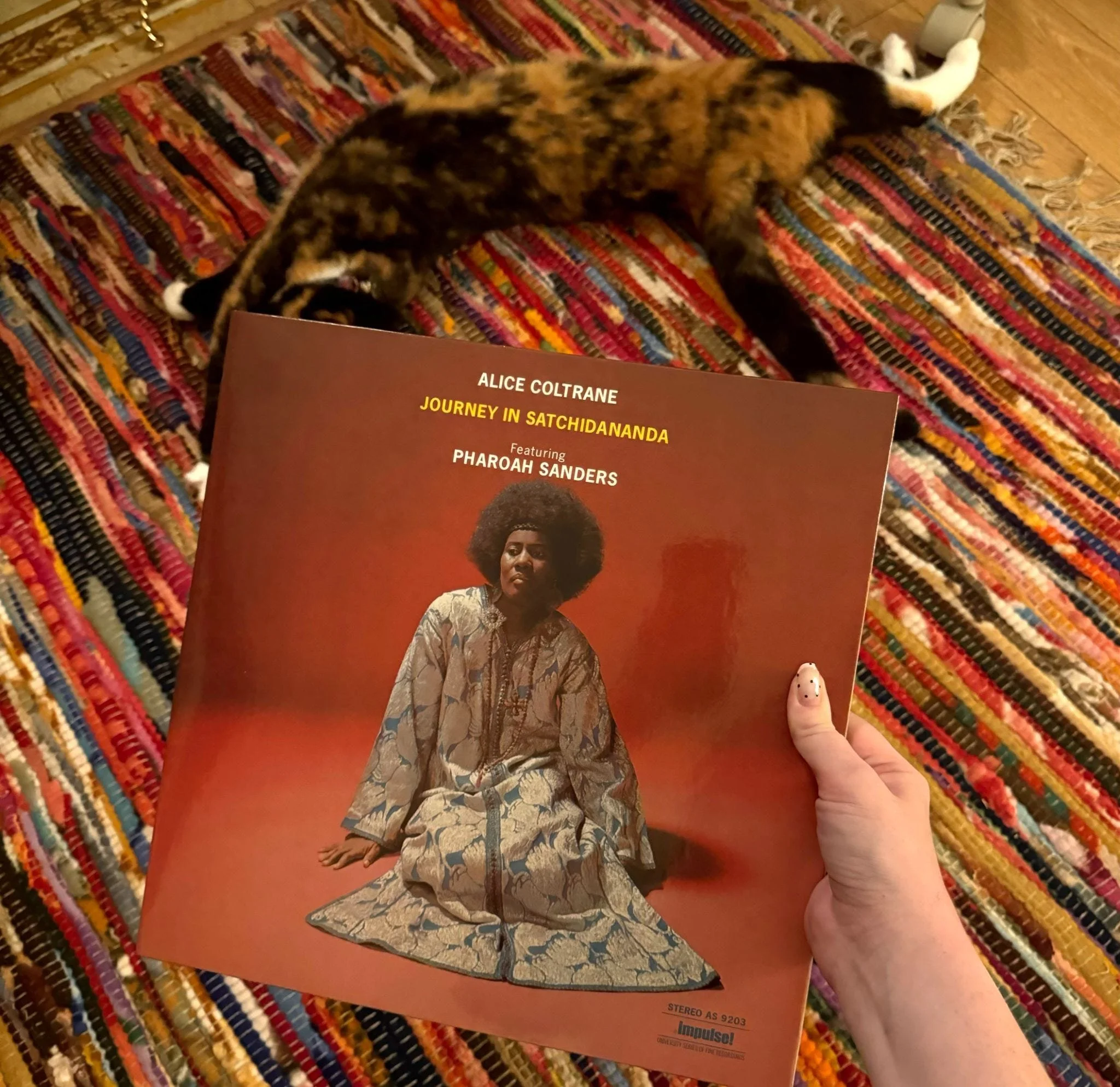 Alice Coltrane’s Journey in Satchidananda vinyl record held above a colorful rug, with a cat lying in the background.