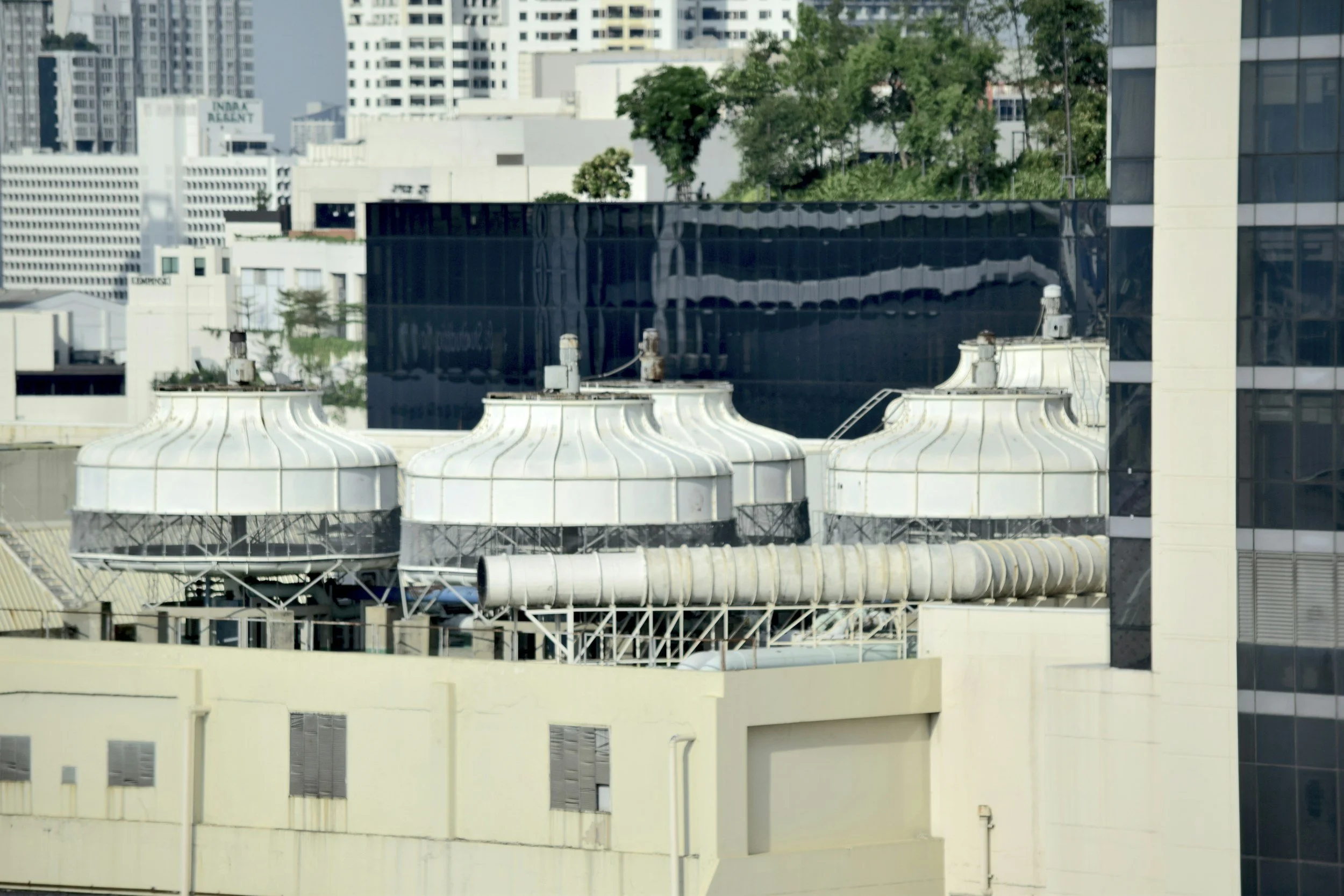 Rooftop view of three white industrial cooling towers on a building in an urban cityscape with high-rise buildings and green trees in the background.