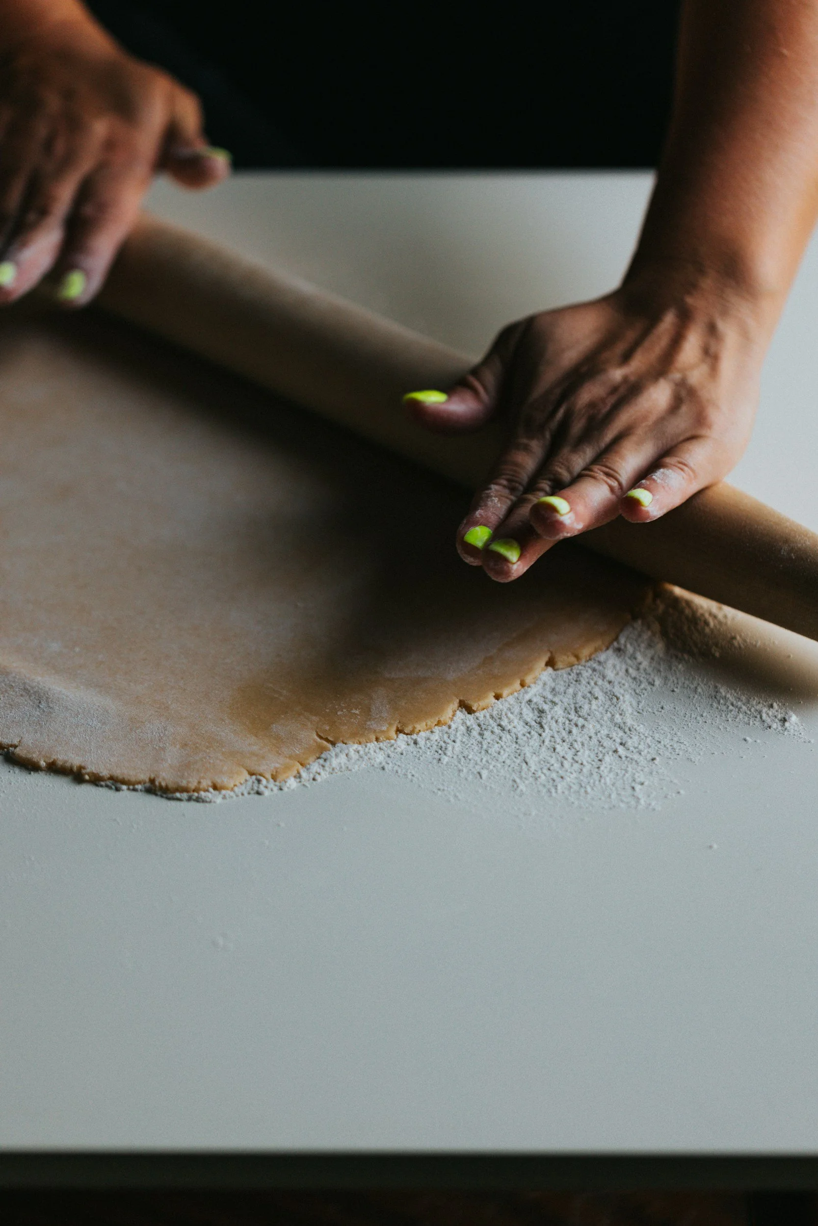 Person rolling out cookie dough with a rolling pin on a white surface, with flour sprinkled underneath.