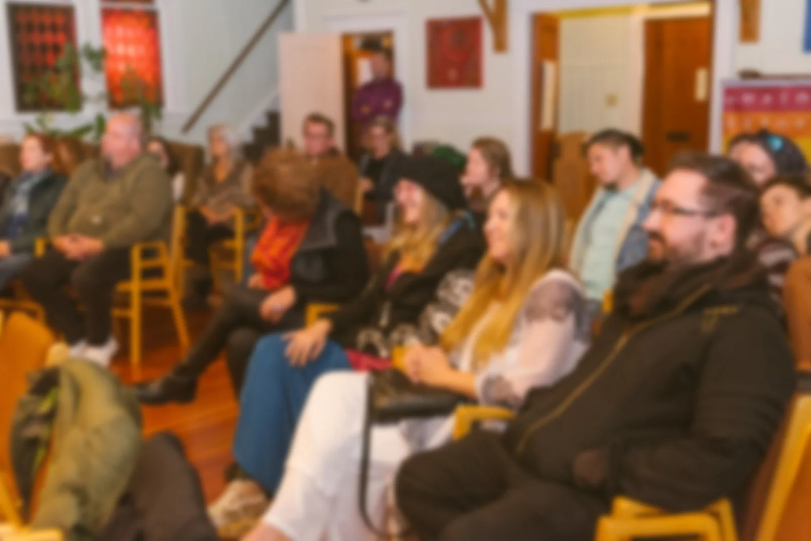 Group of people standing and sitting in a room with large windows, which is at VTC (Victoria Truth Centre) some engaged in conversation, others observing or sitting, during a community event which included live music and connection.