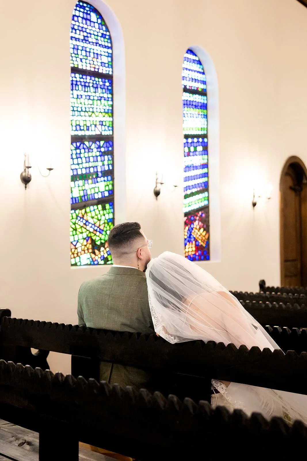 A couple at a wedding ceremony seated in a church with stained glass windows.