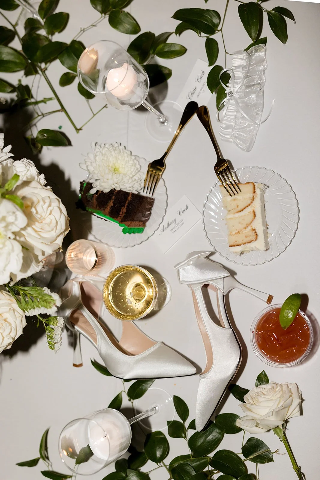Overhead view of a dining table with white flowers, two glasses of wine, a glass of iced tea, a wrapped gift, a pair of satin high heels, a cake with a fork, a slice of cake with a fork, a candle, and a place card.