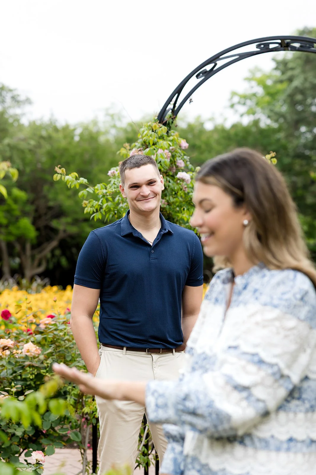 A couple standing in a garden with colorful flowers, with the man smiling and the woman looking down, holding her hands out.