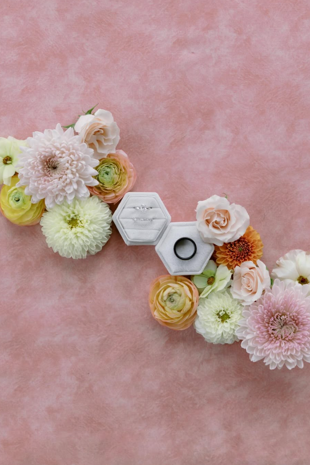 A floral arrangement of pastel-colored flowers surrounds two white jewelry boxes, one containing rings and the other a black band, on a pink textured background.