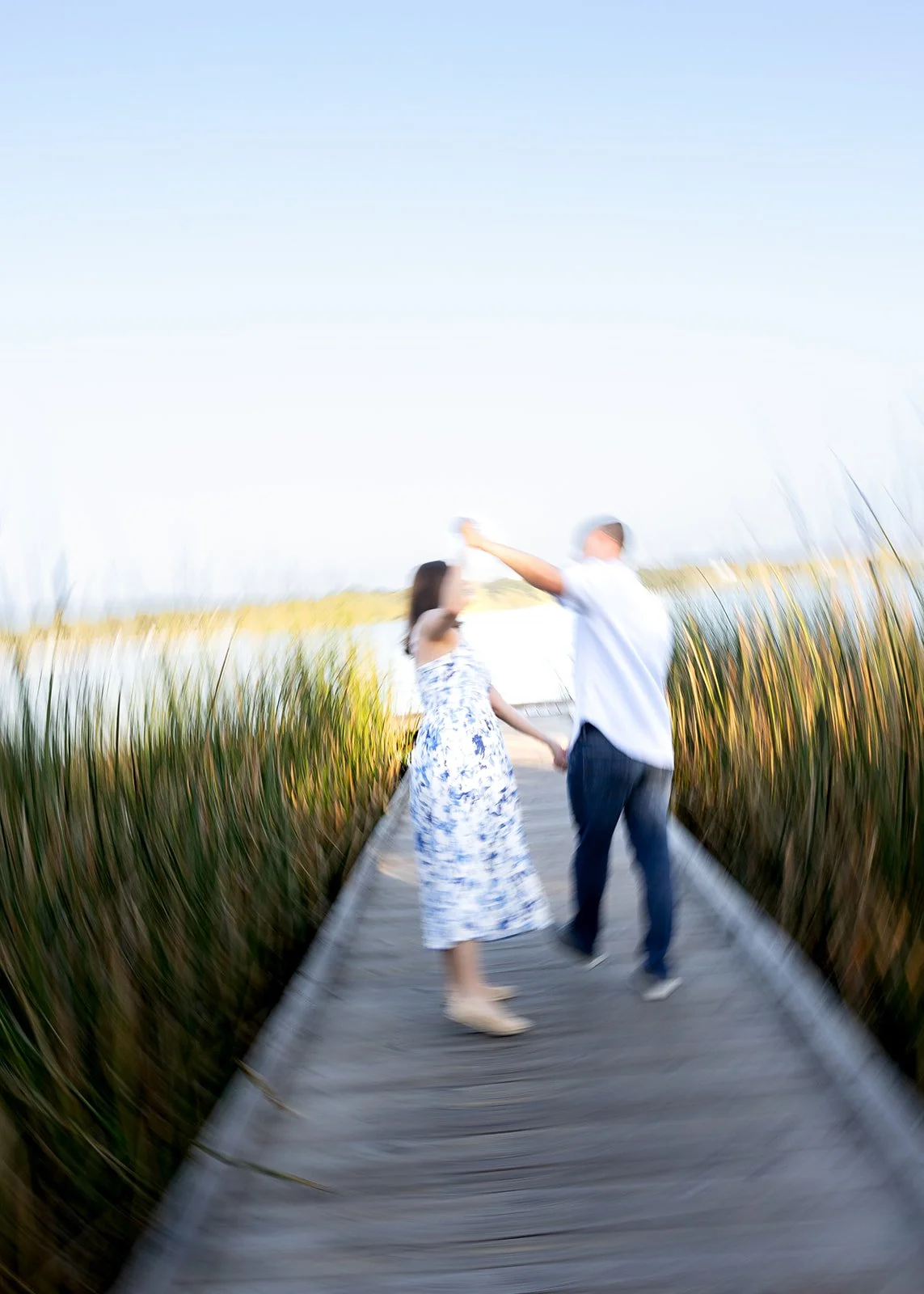 A couple holding hands and dancing on a wooden boardwalk through a marshland with tall grasses, under a clear sky.