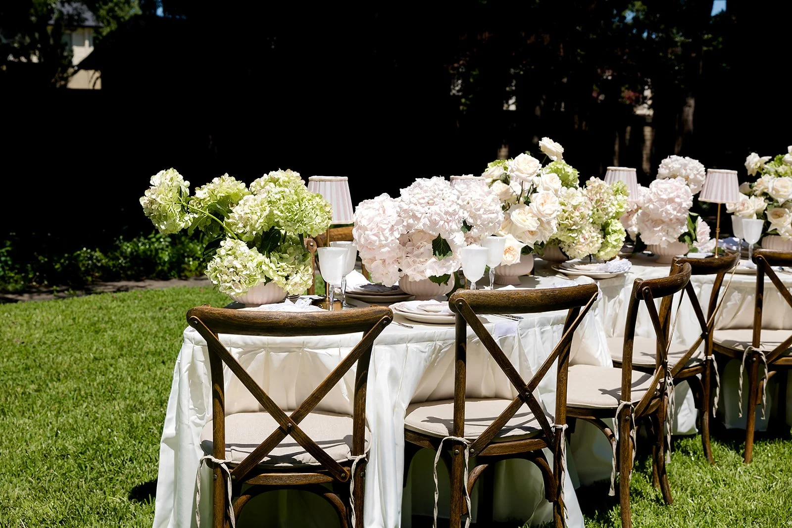 Outdoor wedding reception table at The Dallas Arboretum with white tablecloth, floral centerpieces of white and pale pink hydrangeas and roses, wooden chairs, and small table lamps, on a grassy lawn.