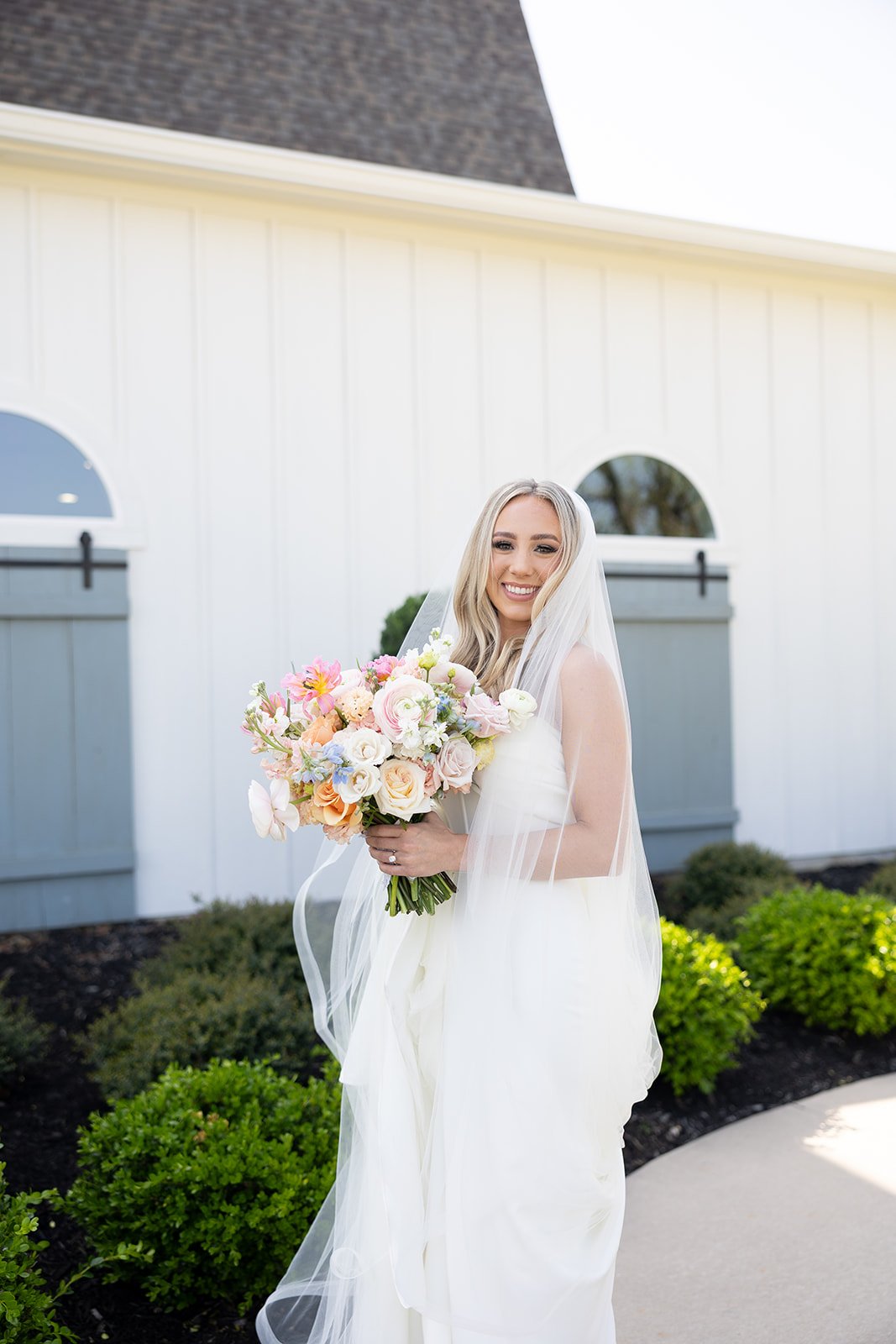 A smiling bride in a white wedding dress at The French Farmhouse and veil holding a bouquet of pink, white, and peach flowers outside near a white building and greenery.