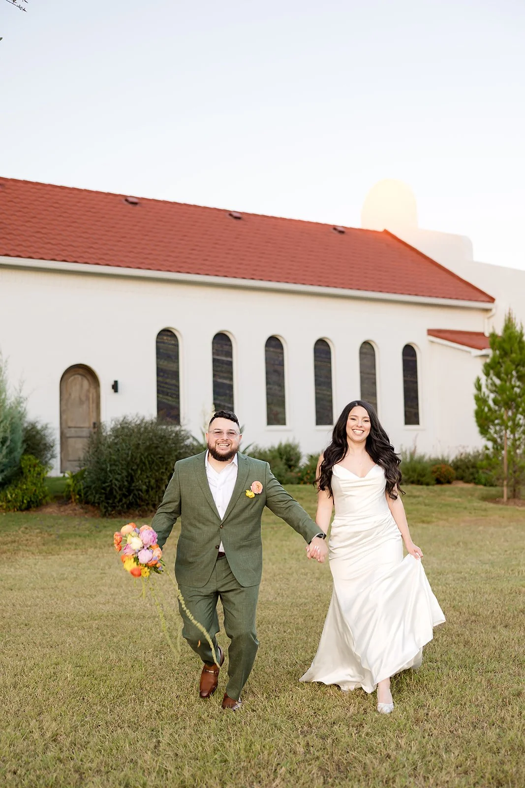 A smiling couple in wedding attire walking hand in hand on a grassy area in front of a white church with a red roof during sunset. The man is wearing a green suit and holding a colorful bouquet, while the woman is dressed in a white wedding gown.