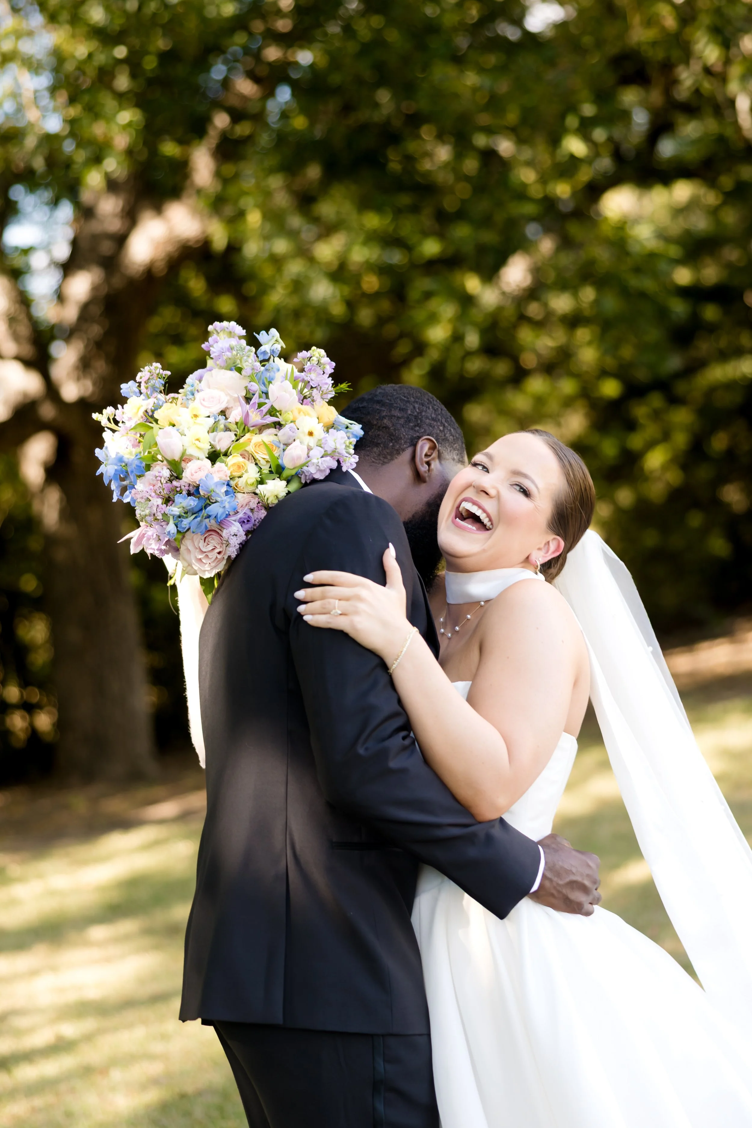 A joyful bride and groom at The Olana in wedding attire embracing outdoors, with the bride laughing and holding a bouquet of pastel-colored flowers, trees in the background.