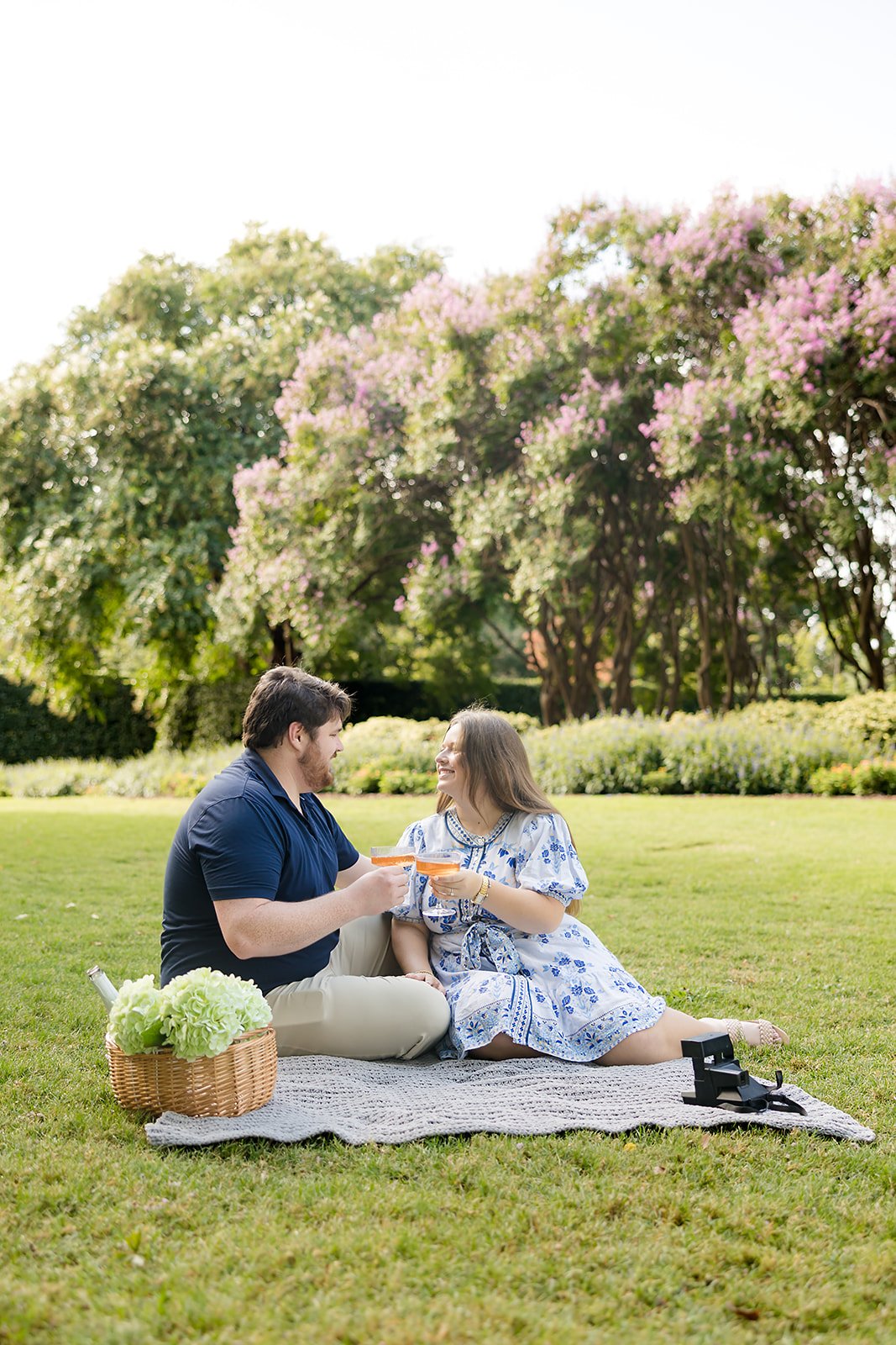 A man and a woman sitting on a blanket on a grassy field, enjoying drinks together in a park with trees and pink blossoms in the background.