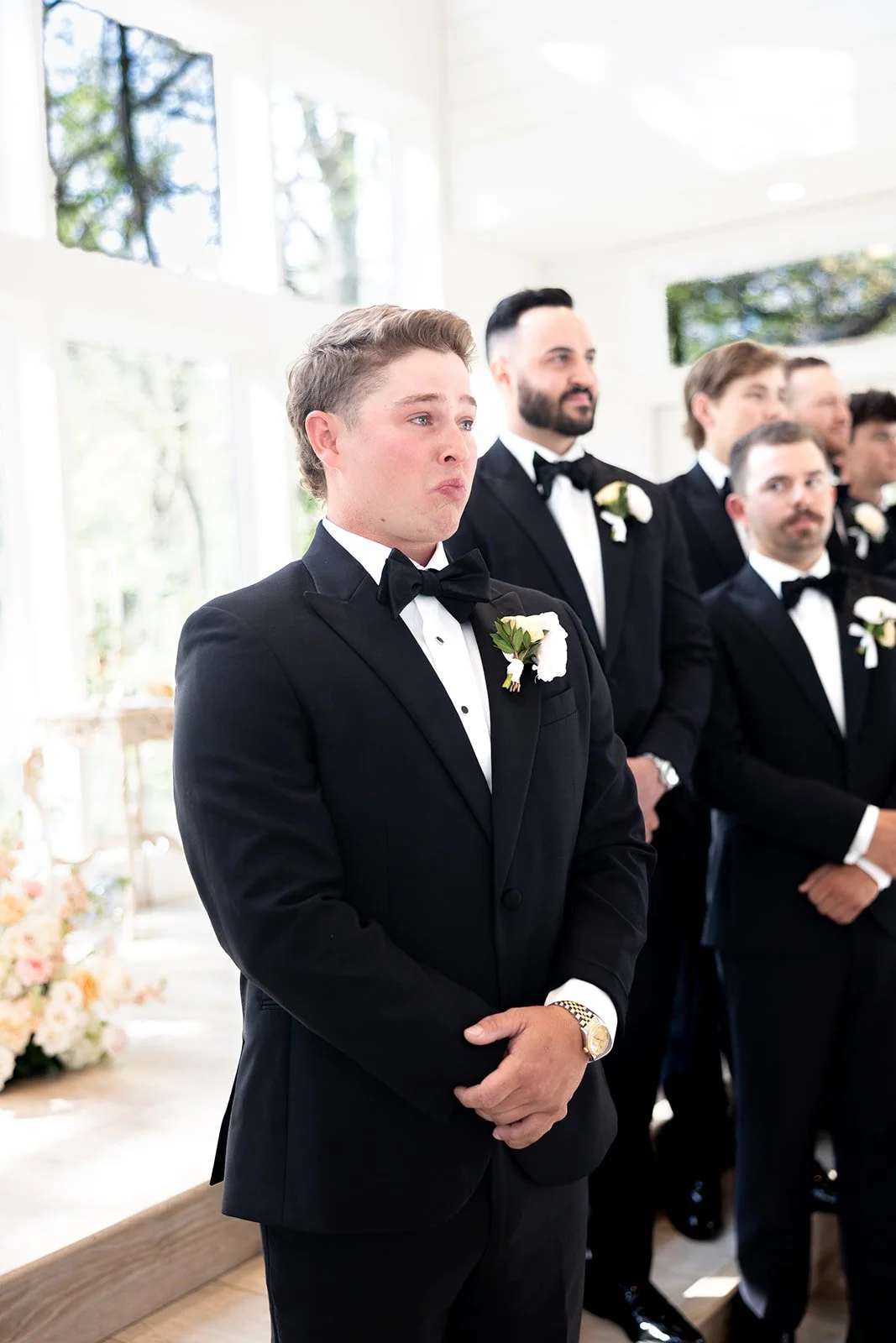 A young man in a black tuxedo with a bow tie and boutonniere stands with clasped hands during a wedding ceremony, surrounded by other men in similar attire inside a bright room with large windows and natural light.