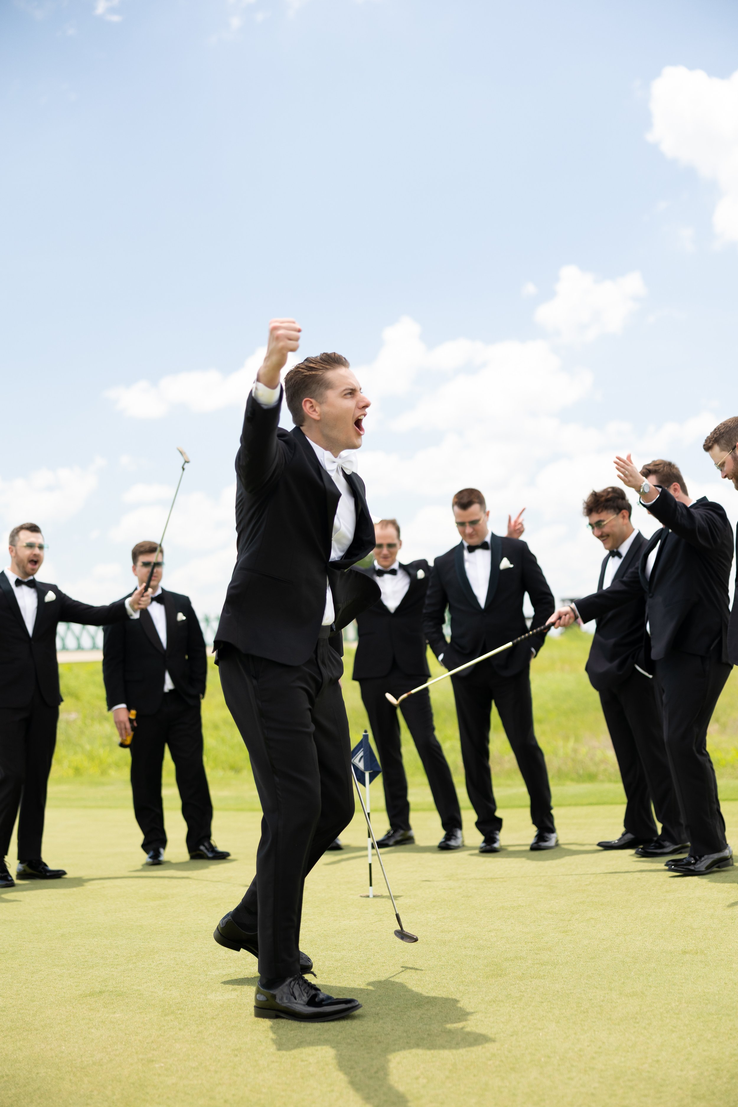 Group of men in tuxedos celebrating on a golf course with one man holding a golf club and raising his fist. Golf Wedding