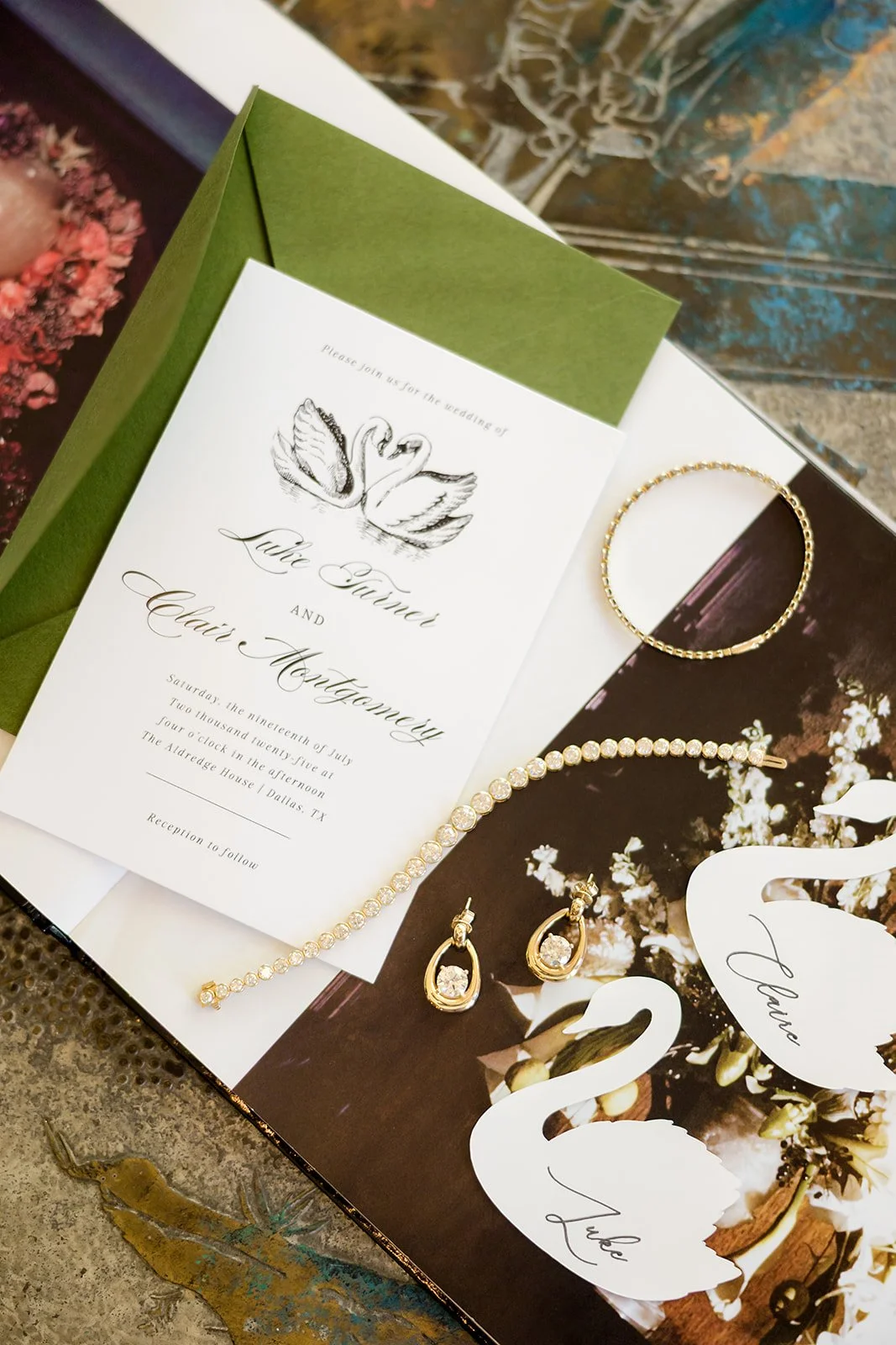 Wedding invitation with black swan and couple's names, Louie Turner and Claire Montgomery, along with a gold bracelet, gold earrings with clear stones, and swan-shaped thank you cards on a decorative background.