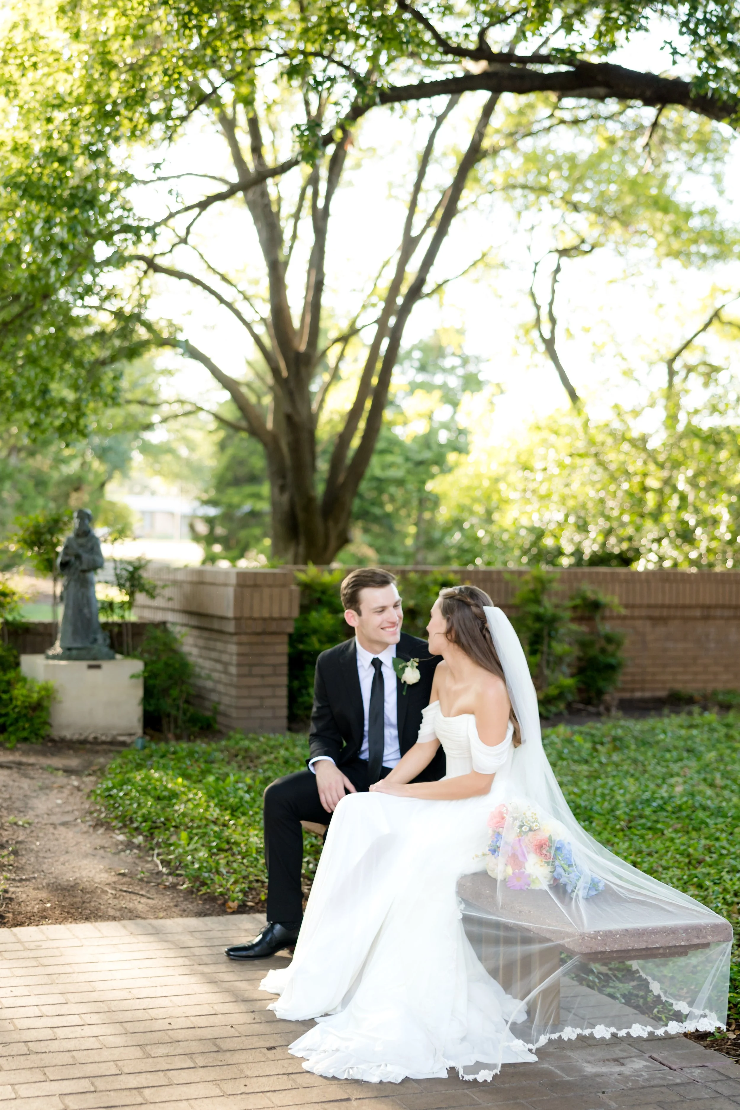 A bride and groom sitting on a bench outdoors, smiling at each other, with trees and a brick wall in the background during daytime.
