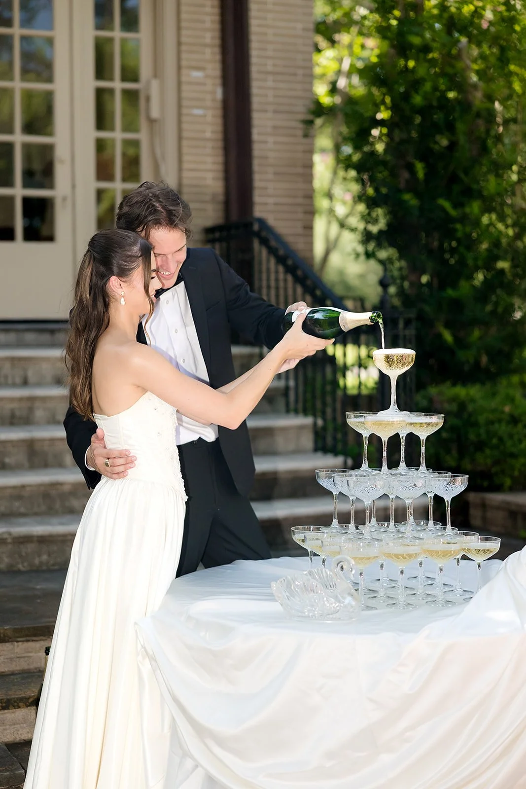 A groom pours champagne into a tower of glasses on a table outside during a celebration.
