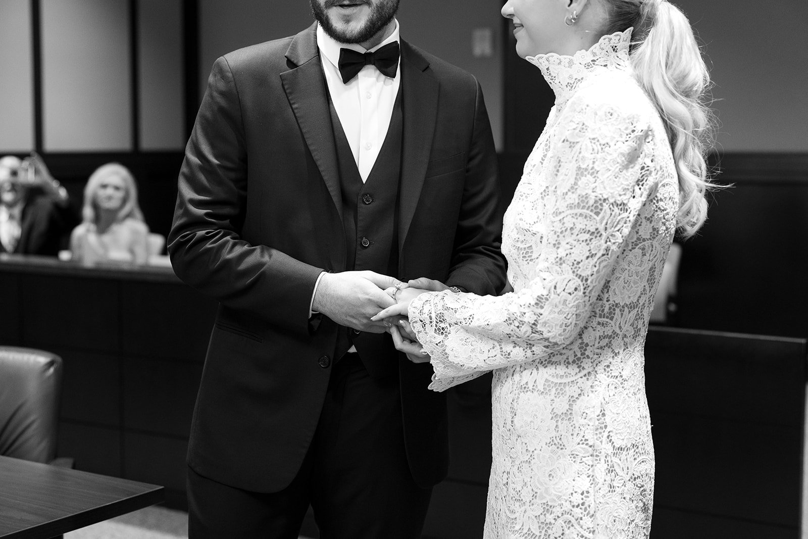 Black and white photo of a wedding ceremony showing a groom and bride holding hands, with the groom wearing a tuxedo and the bride in a lace wedding dress.