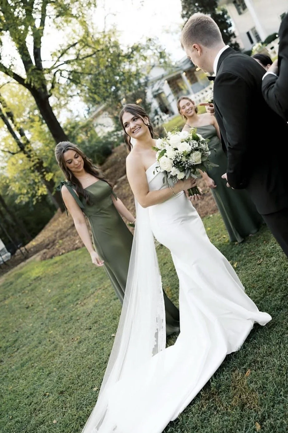 Wedding ceremony outdoors with bride in a white gown holding a bouquet of white and green flowers, groom in a black suit, and bridesmaids in green dresses, trees, and a house in the background.