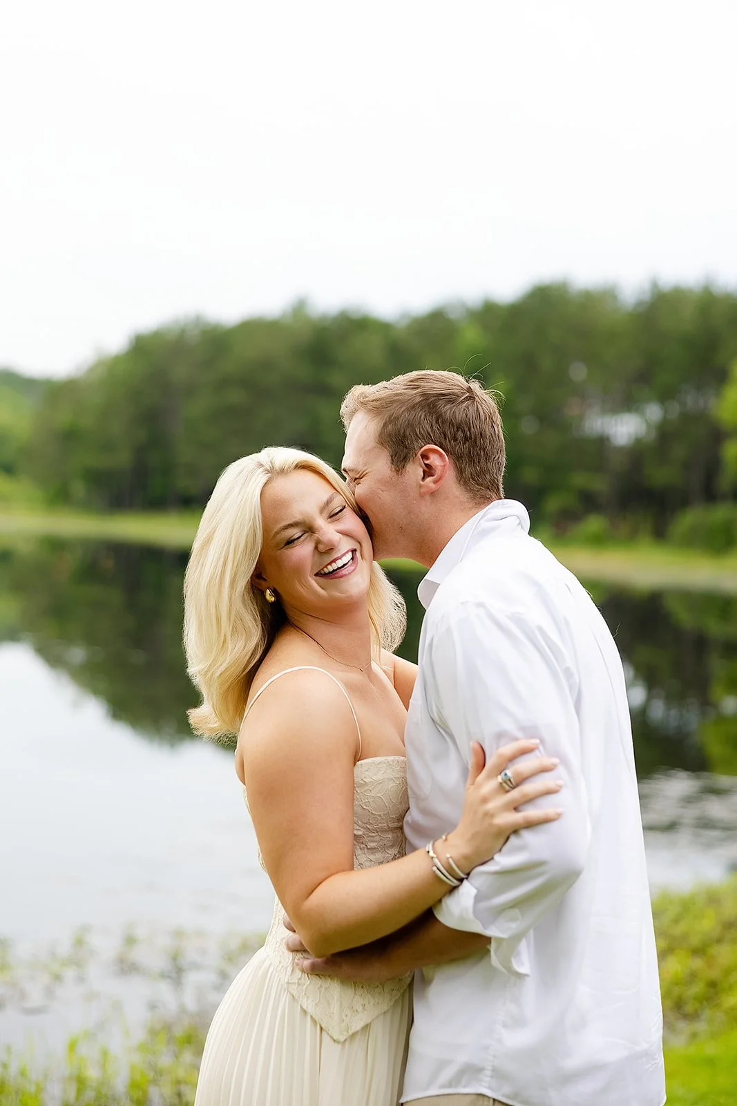 A couple near a lake, the woman with blonde hair wearing a light dress, the man with short hair in a white shirt, sharing a joyful moment, the man kissing the woman's cheek, in a lush green outdoor setting.