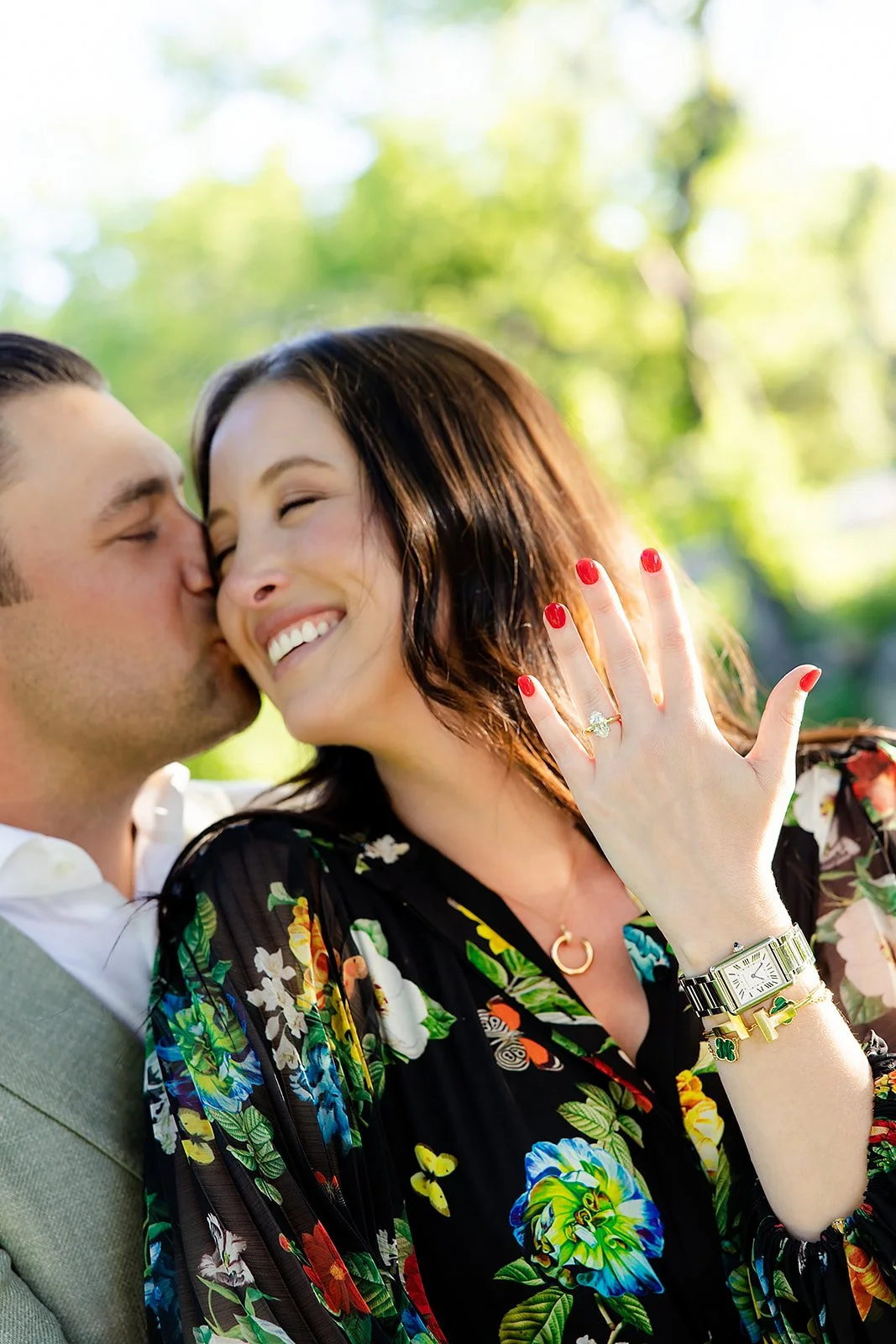 A woman showing off a ring on her finger while smiling and a man kissing her cheek outdoors on a sunny day with green trees in the background.