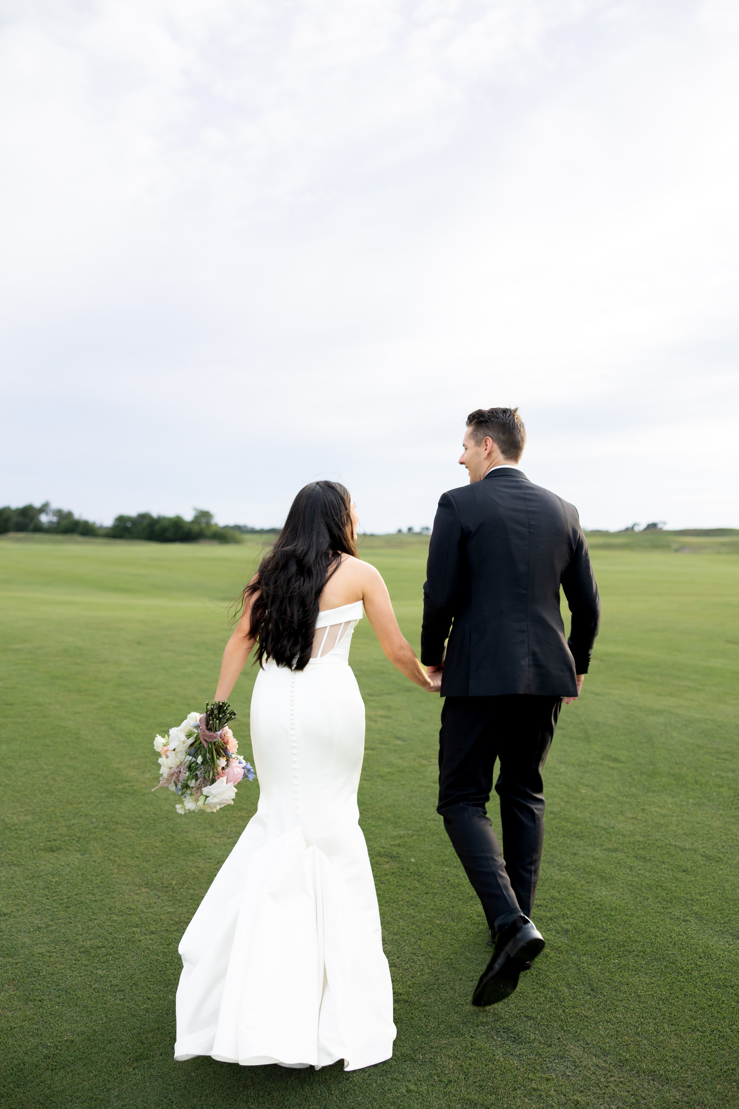 A newlywed couple walking hand in hand on a grassy field, with the bride holding a bouquet of flowers and wearing a white wedding dress, while the groom is dressed in a black suit. The sky is partly cloudy.