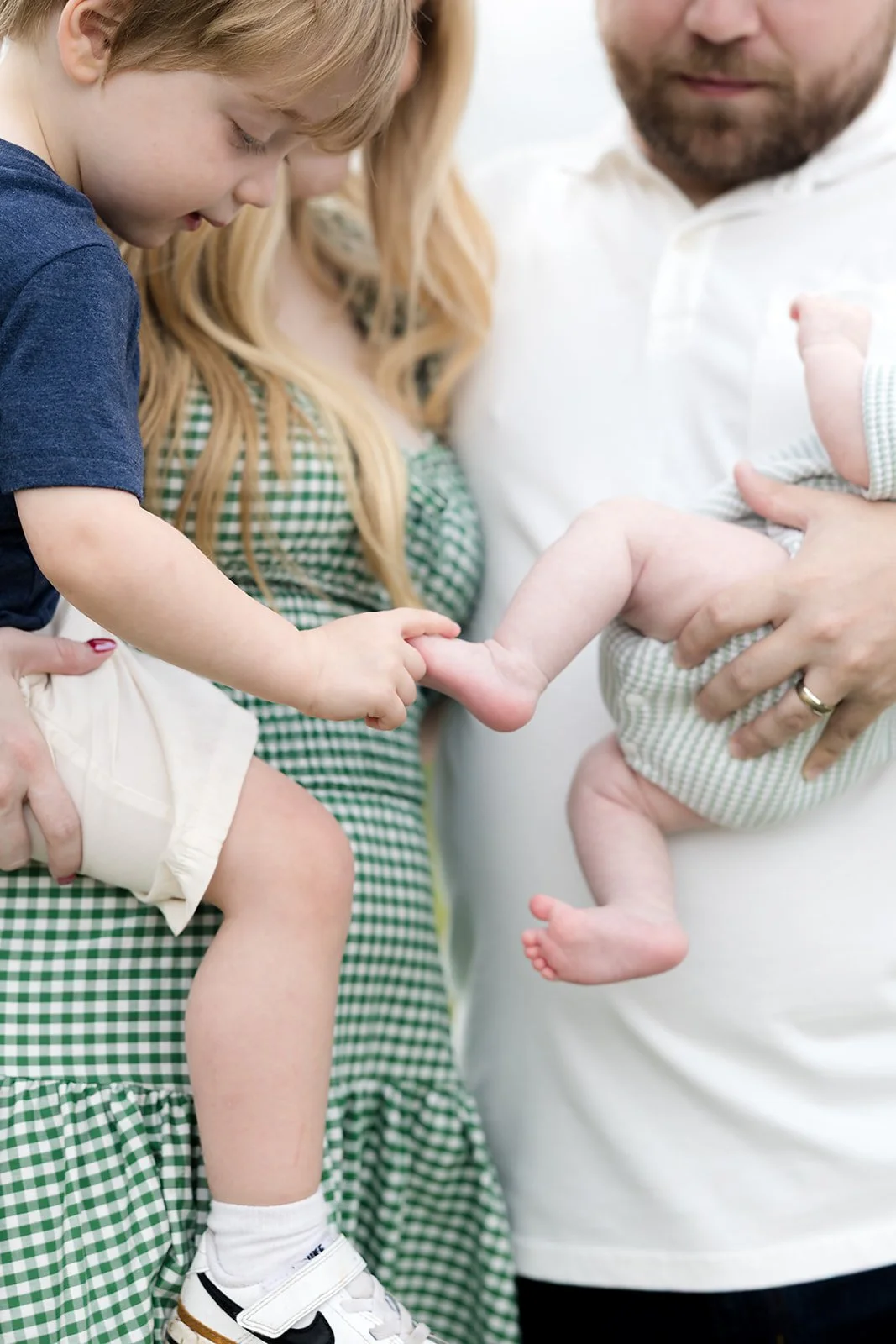 A young boy with blonde hair touches the foot of a baby held by an adult man, with a woman in a green checkered dress nearby.