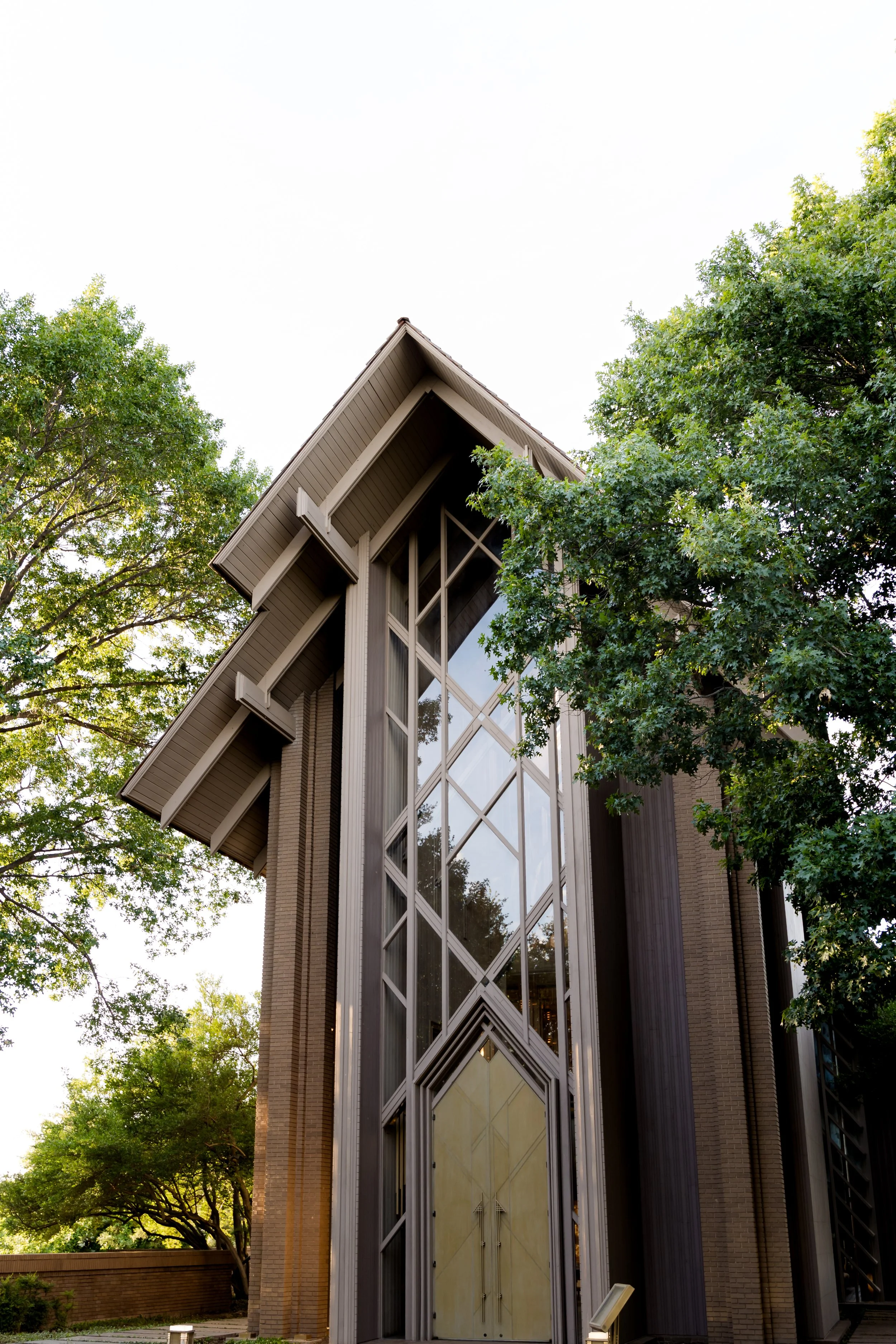Modern church building with large glass windows and metal framework, surrounded by green trees.